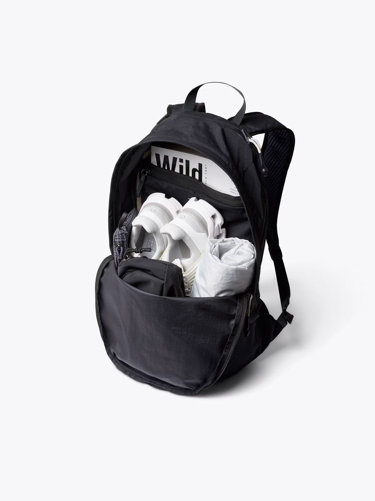 The Bellroy Lite Daypack - 20L is shown open, displaying its spacious main compartment packed with a rolled-up white shirt, white sneakers, a small black pouch, patterned fabric roll, and a WILD magazine.