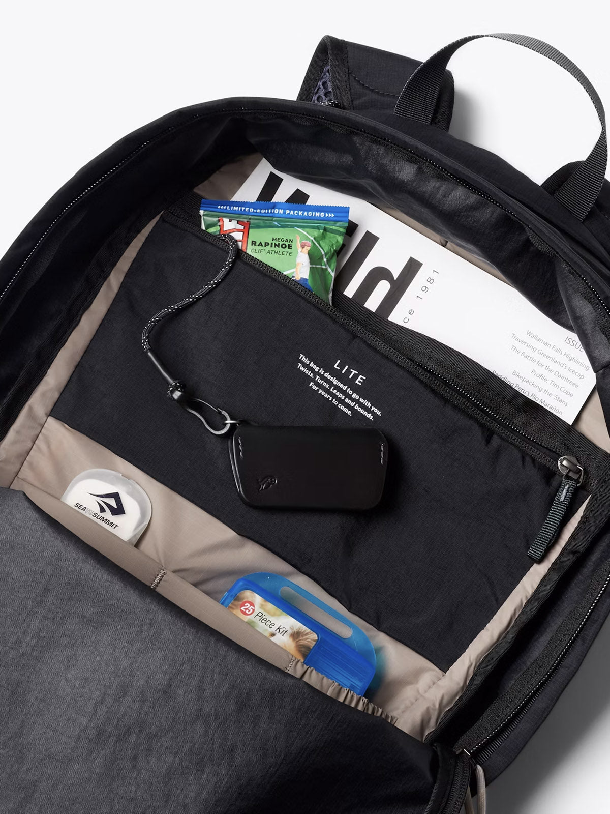 The Bellroy Lite Daypack - 20L is open to reveal organized compartments holding a magazine, national park pass, wireless earbud case, keychain, blue gum pack, and granola bar.
