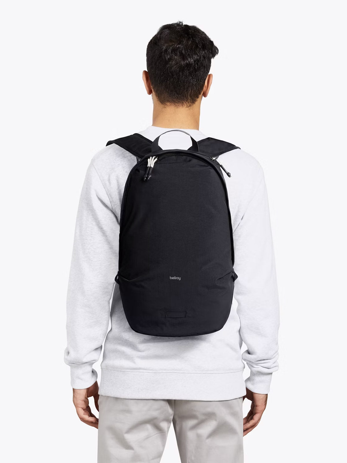 A person is seen from behind carrying the Bellroy Lite Daypack - 20L, featuring white zipper pulls, over a light gray sweatshirt and beige pants against a plain, light background.