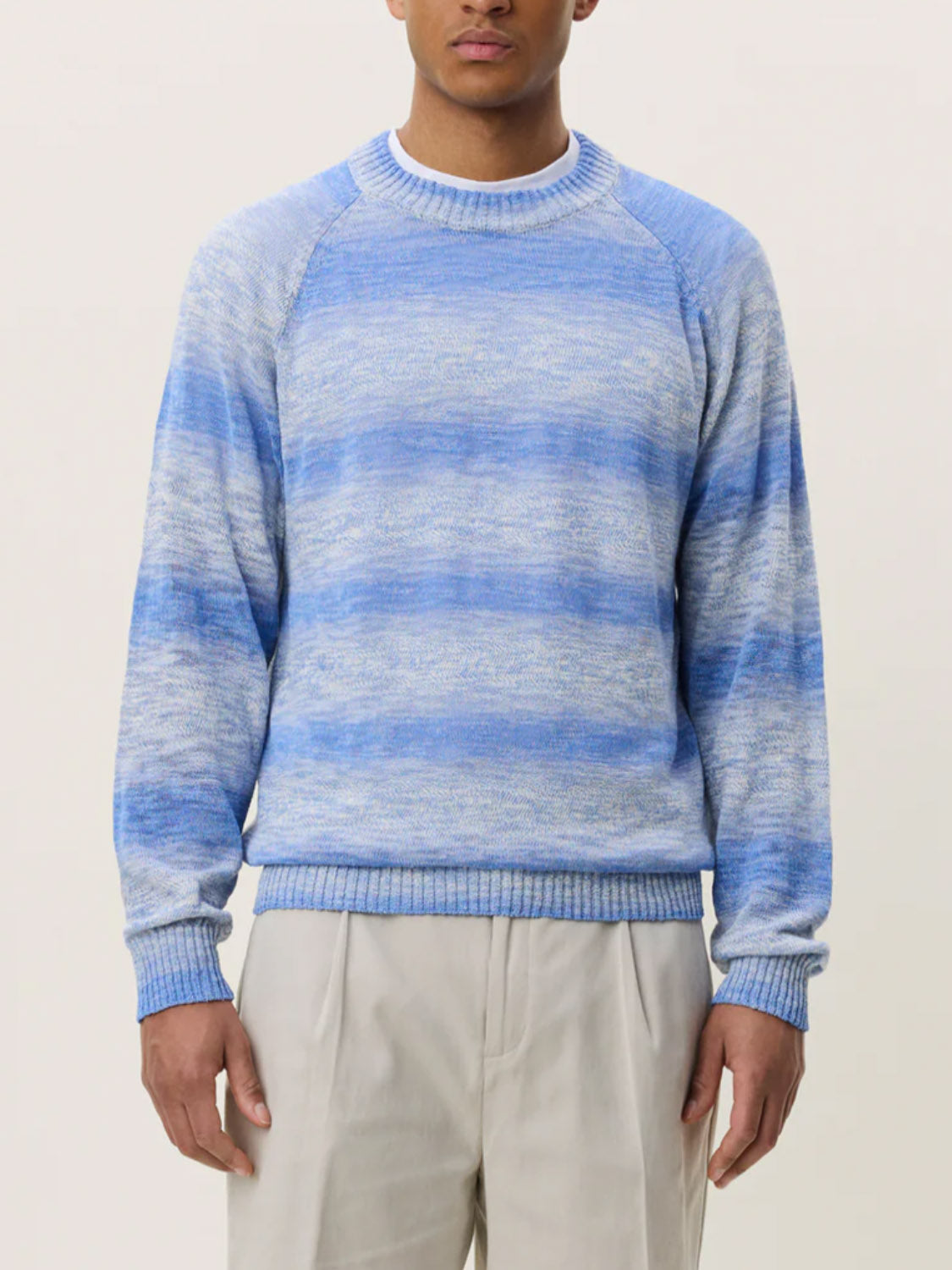 A person wearing the Les Deux Light Gradient Knit—a light blue and white marbled crewneck sweater—paired with light beige pants stands against a plain, light background.