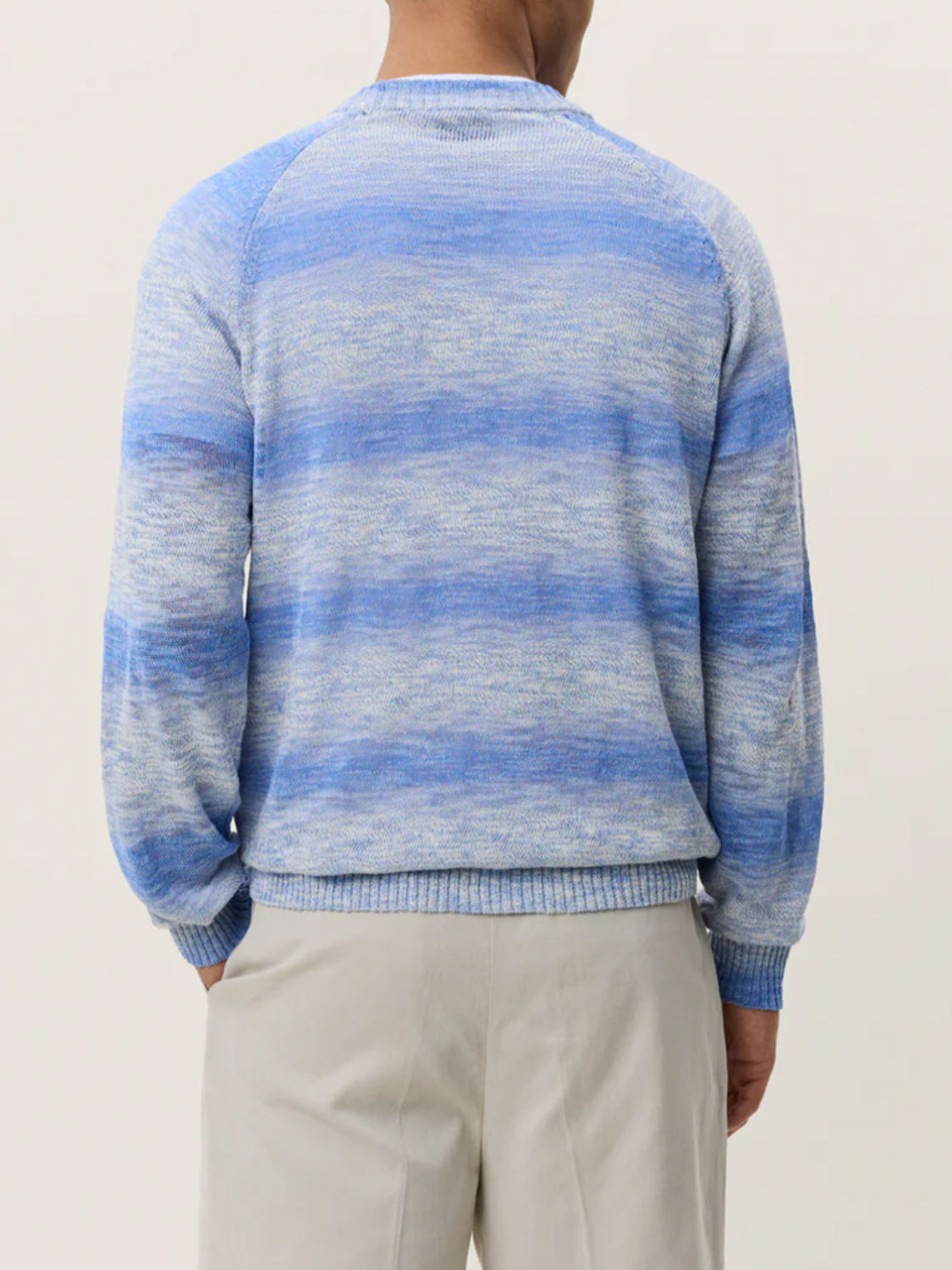 A person is shown from the back against a plain light background wearing the Les Deux Light Gradient Knit sweater in light blue and white stripes paired with light beige pants.