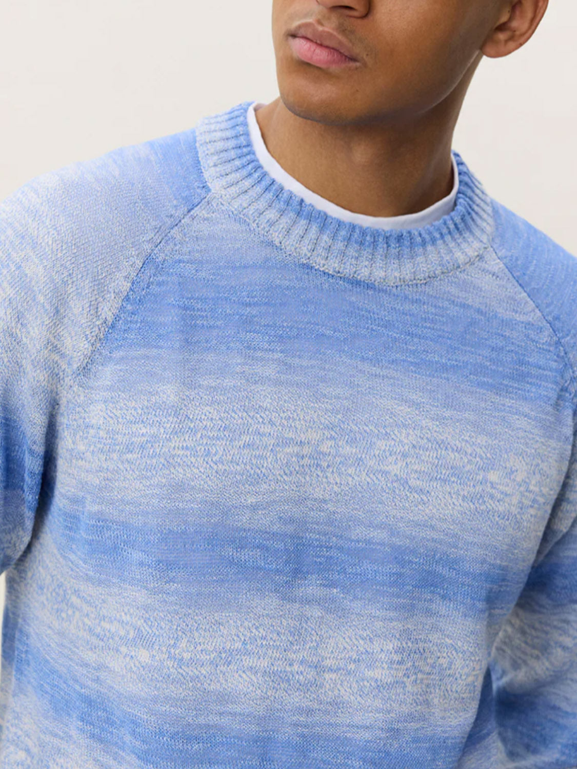 A person wears the Les Deux Light Gradient Knit, a light blue and white cotton-tencel crewneck sweater layered over a white shirt, shown from shoulders to mid-chest with their face partially out of frame against a plain background.
