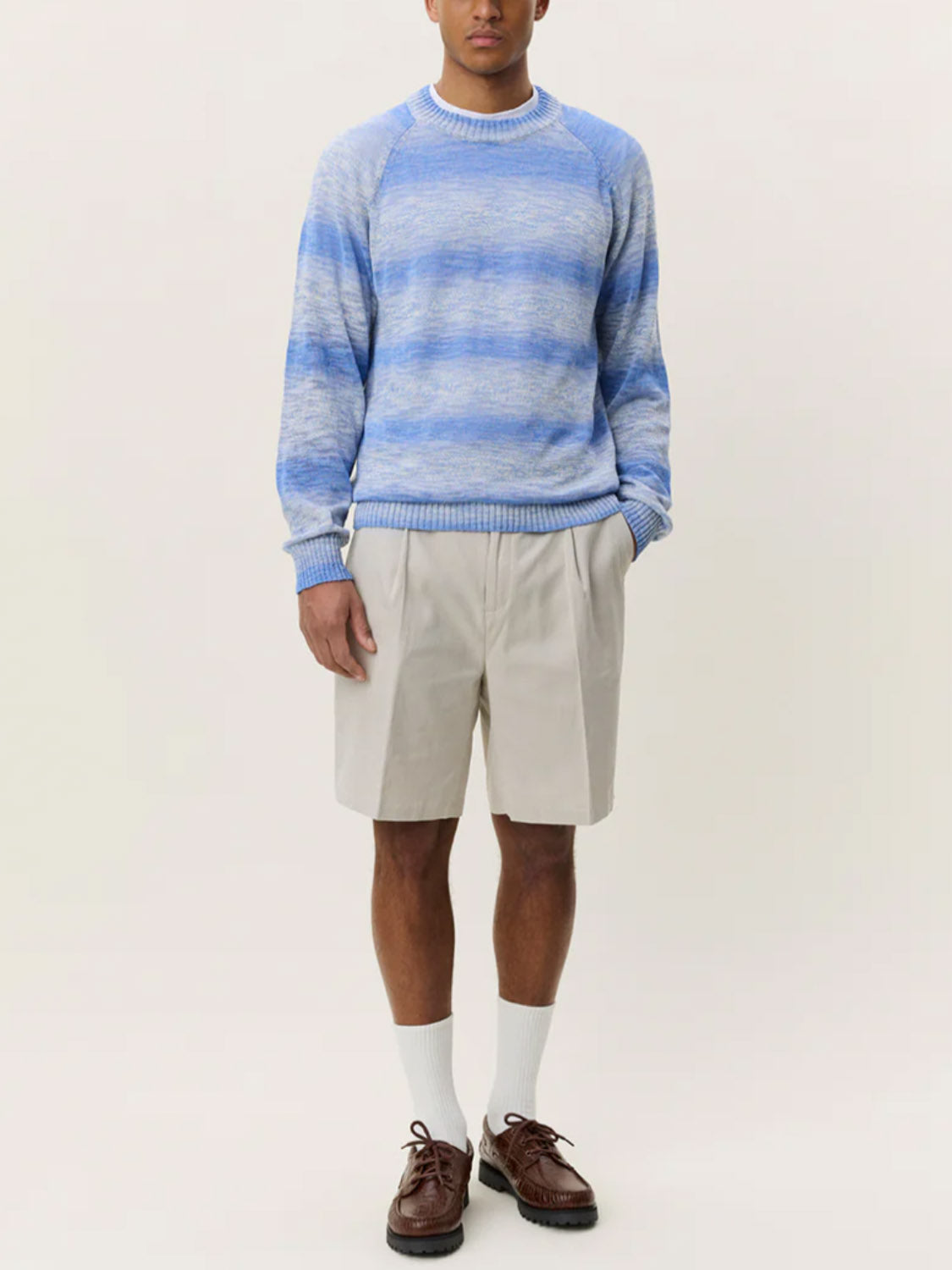 A person stands against a plain light background wearing the Les Deux Light Gradient Knit sweater, beige knee-length shorts, white socks, and brown shoes with hands in their pockets.