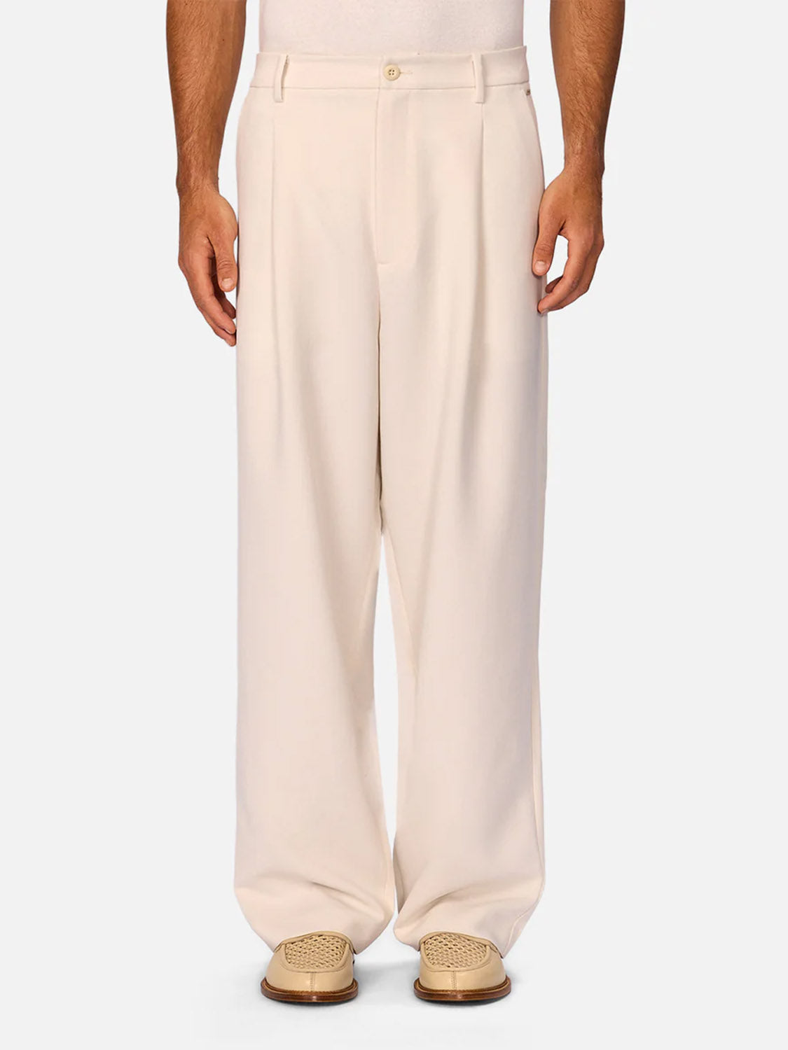Wearing Industrie’s The Light Weight Lusso Pant—high-waisted, wide-leg cream trousers with pleats—paired with light beige woven loafers, a person stands against a plain white background.