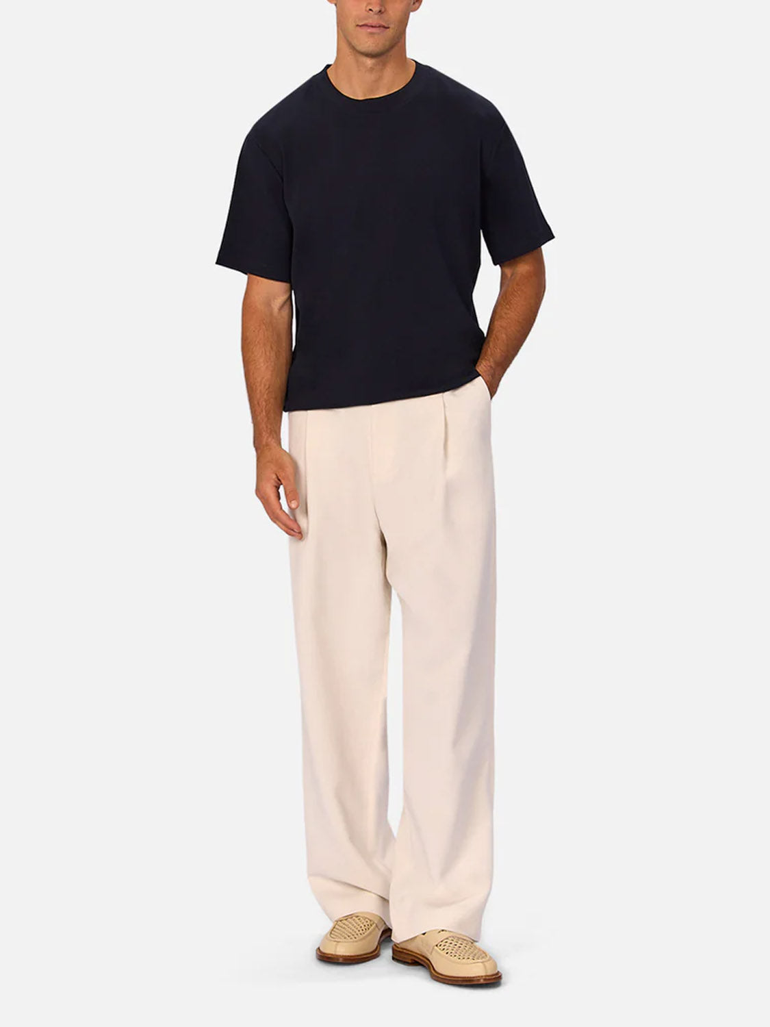 A man stands with his hands in his pockets, wearing a plain black short-sleeve t-shirt, beige woven shoes, and Industrie’s The Lightweight Lusso Pant—wide leg and pleated—against a plain light background.