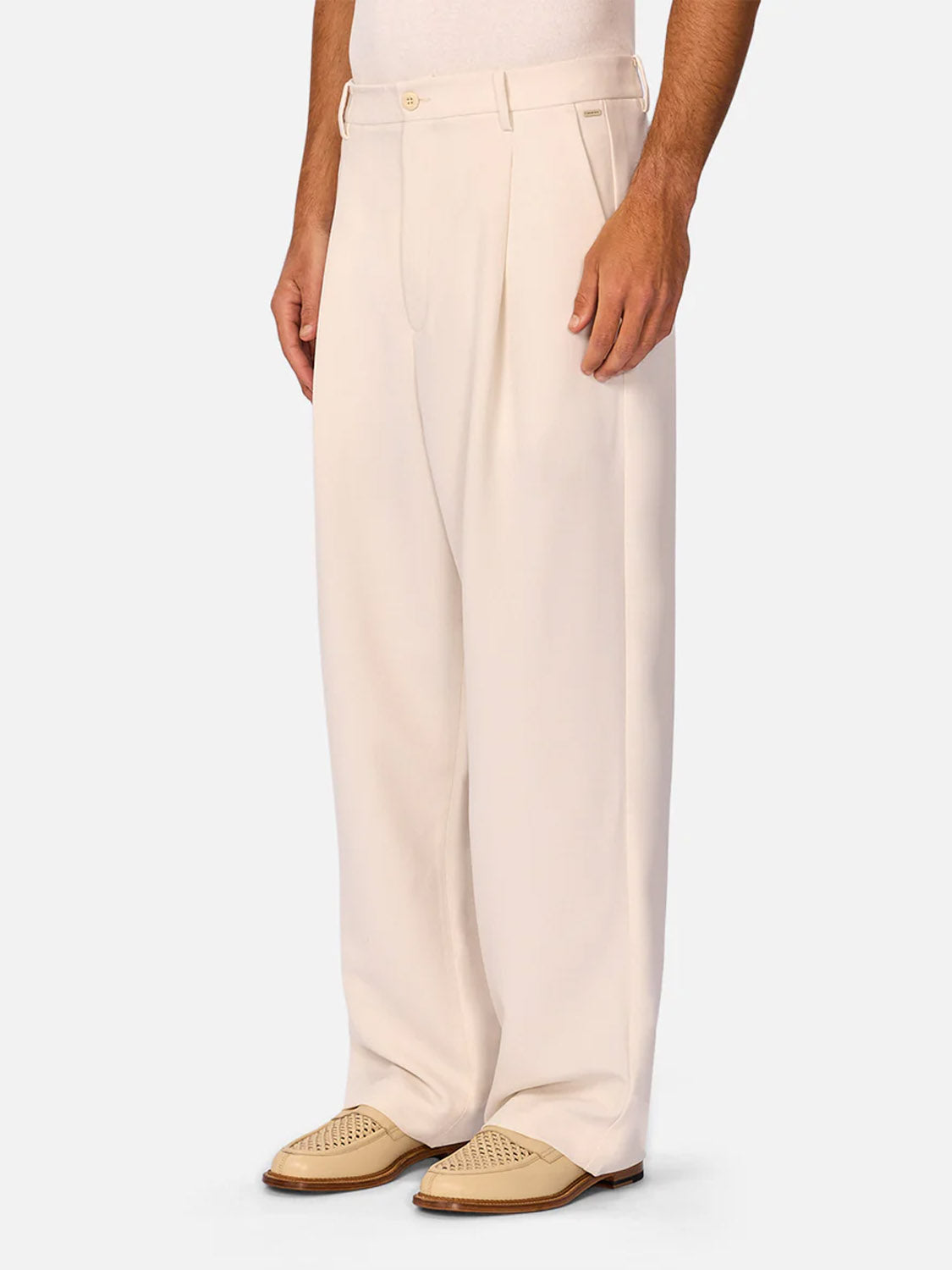 Someone stands against a plain light background wearing Industrie’s The Lightweight Lusso Pant, featuring wide legs, a pleated waistline, and beige button, paired with tan woven leather dress shoes for a refined modern tailored look.