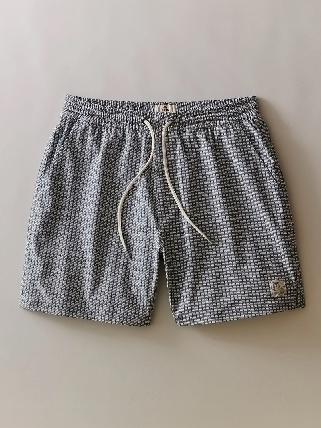 The Marine Layer Lined Sport and Swim Short 6" features a gray and white checkered pattern, elastic waistband with white drawstring, lower left leg patch, and a comfy boxer brief liner for quick-dry performance. Shown flat.