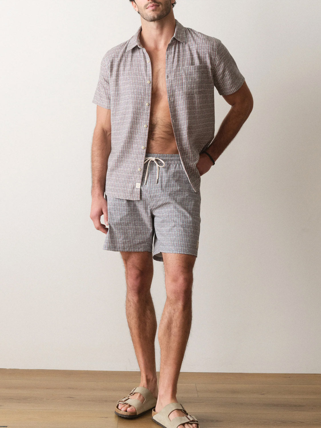 A man indoors wears Marine Layer's Lined Sport and Swim Short 6" and matching shirt—both in checkered print, featuring quick-dry fabric and a stretchy boxer brief liner. He pairs them with beige sandals, no undershirt, against a plain backdrop with wooden flooring.