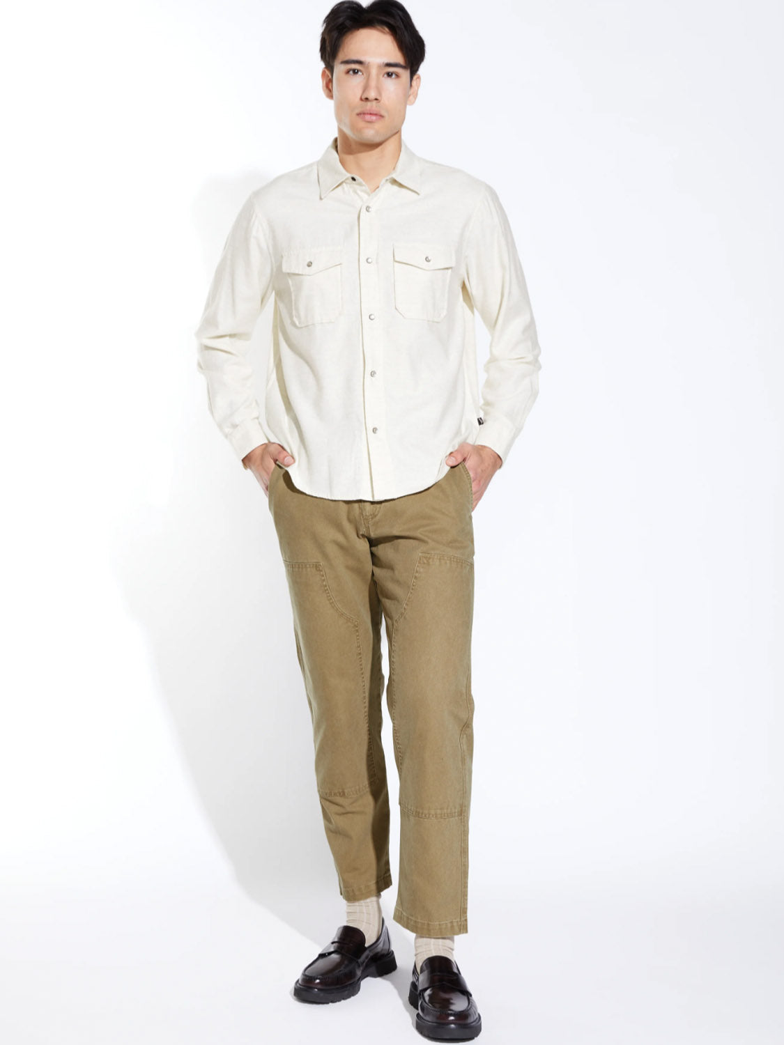 A man stands against a white background wearing the Civil Society Linwood Relaxed Fit Western Shirt in light beige, paired with olive green pants, white socks, and dark brown loafers. His hands are in his pockets and he looks forward.