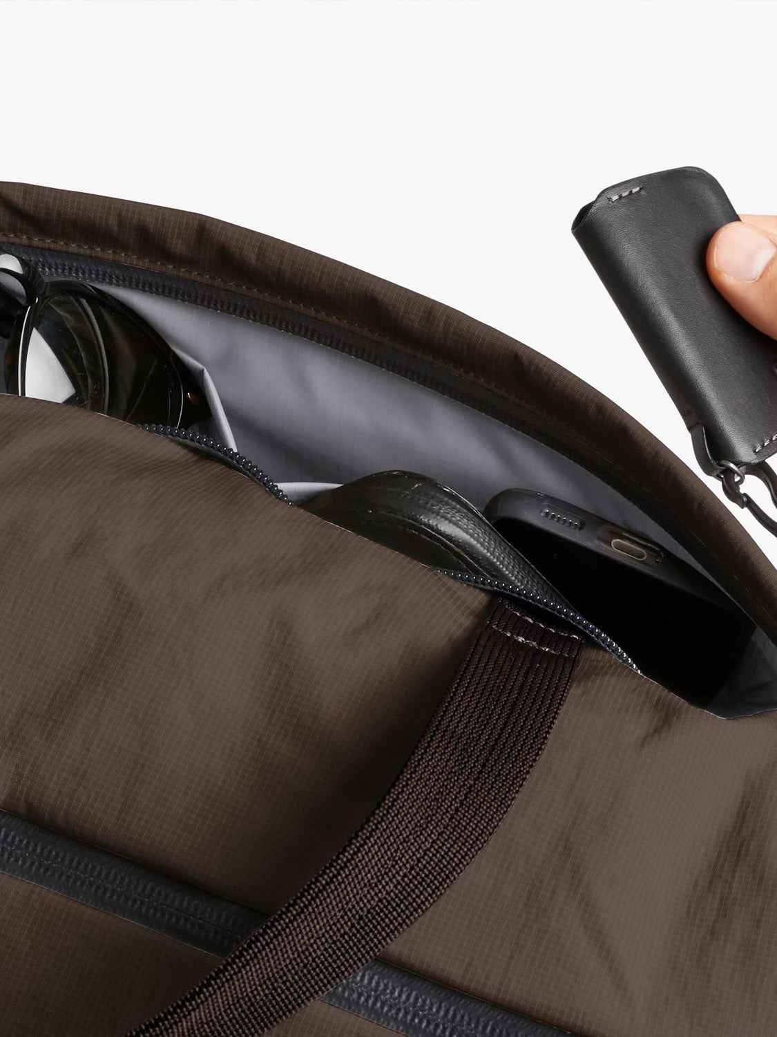 A close-up of a partially open Bellroy Lite Duffle Bag - 30L reveals sunglasses, a smartphone, and more inside, while a hand holds a black key case above the bag.