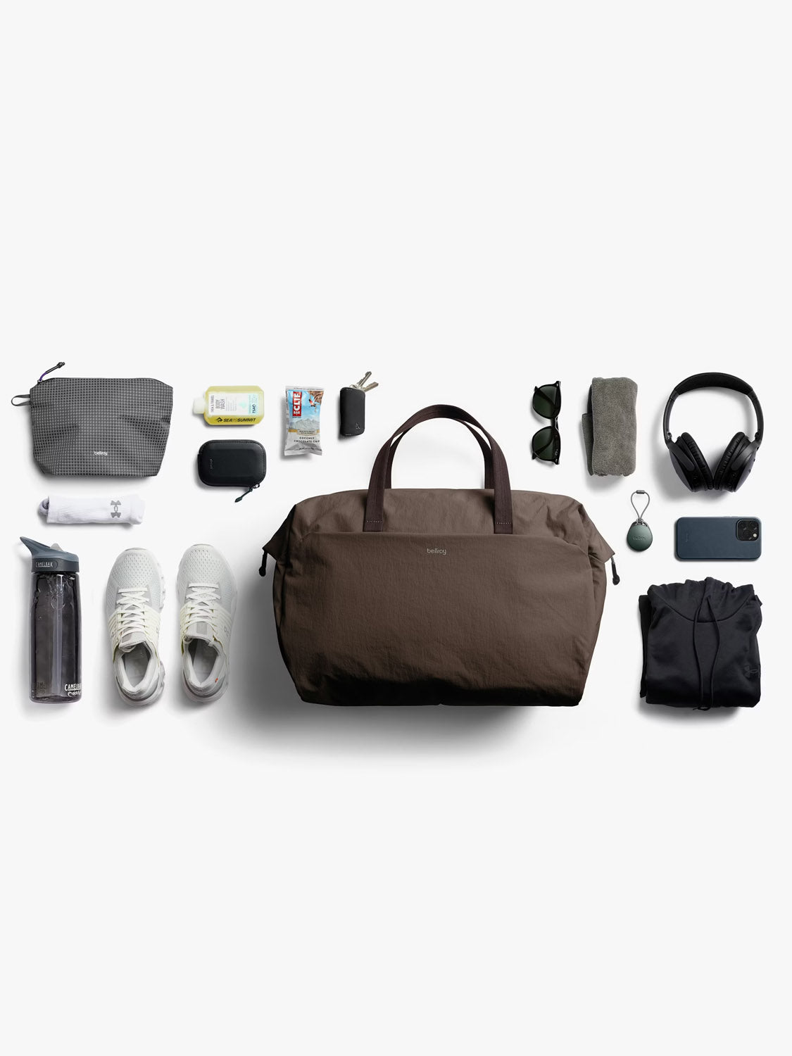 The Bellroy Lite Duffle Bag - 30L is displayed on a white background, surrounded by travel essentials such as sneakers, water bottle, pouch, tissues, earbuds, keys, charger, sunglasses, towel, headphones, shirt, and smartphone.