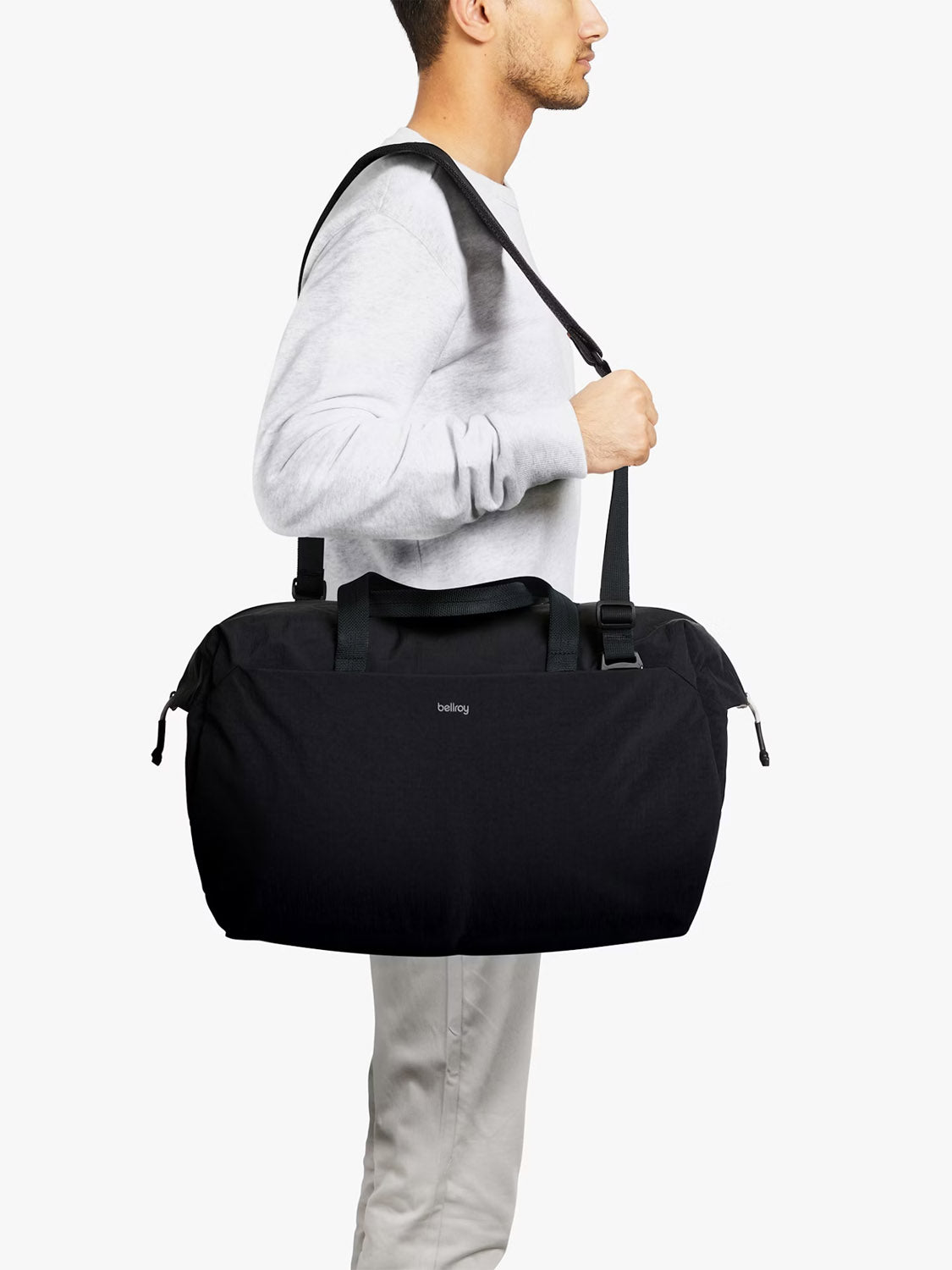 A person in a light gray sweatshirt and beige pants carries the Bellroy Lite Duffle Bag - 30L, featuring both handles and a shoulder strap, standing against a plain white background.