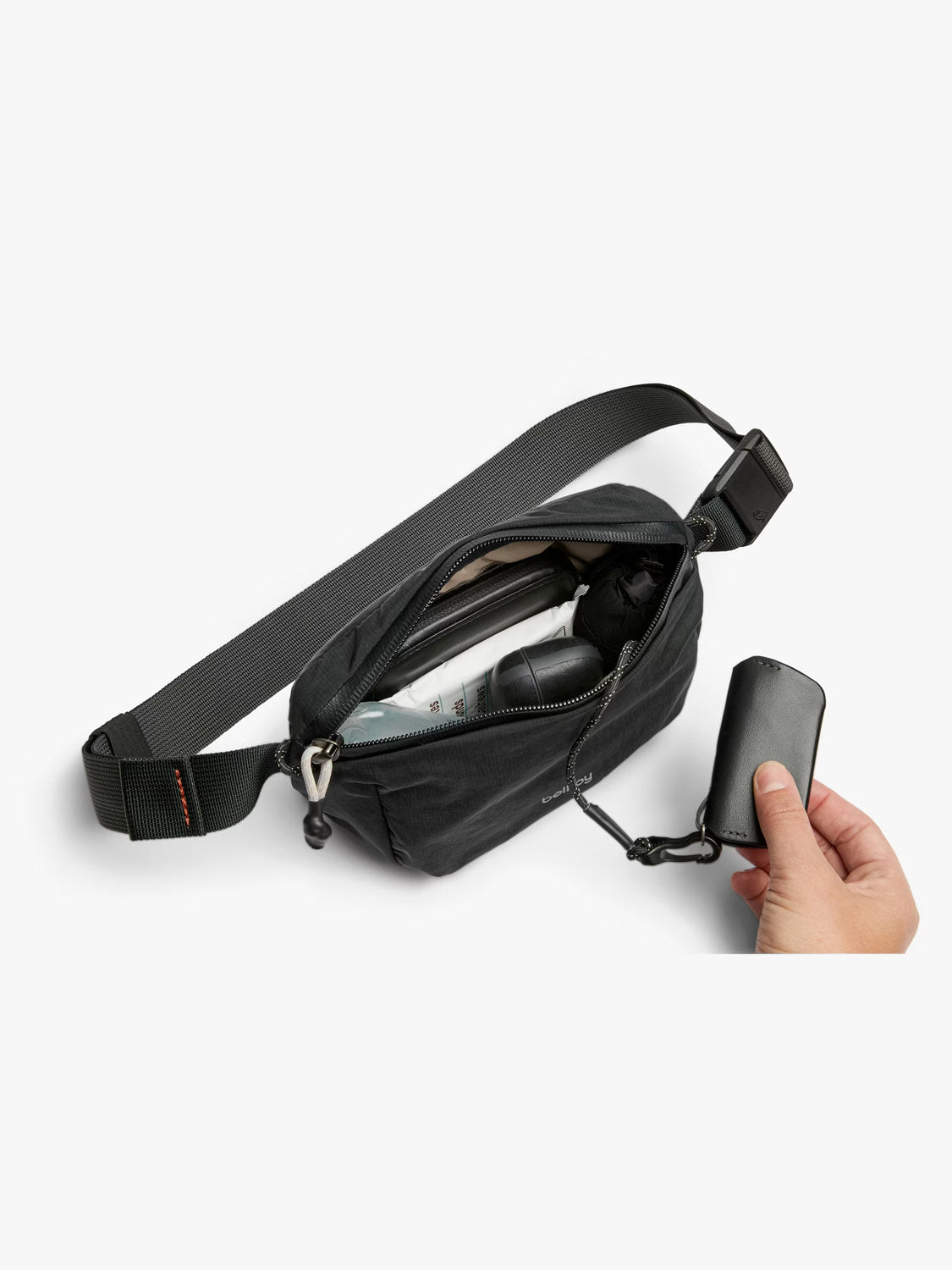 A hand holds a small key case attached to the Bellroy Lite Belt Bag in black, unzipped to reveal neatly organized items inside—such as a pen, a small bottle, and a phone.