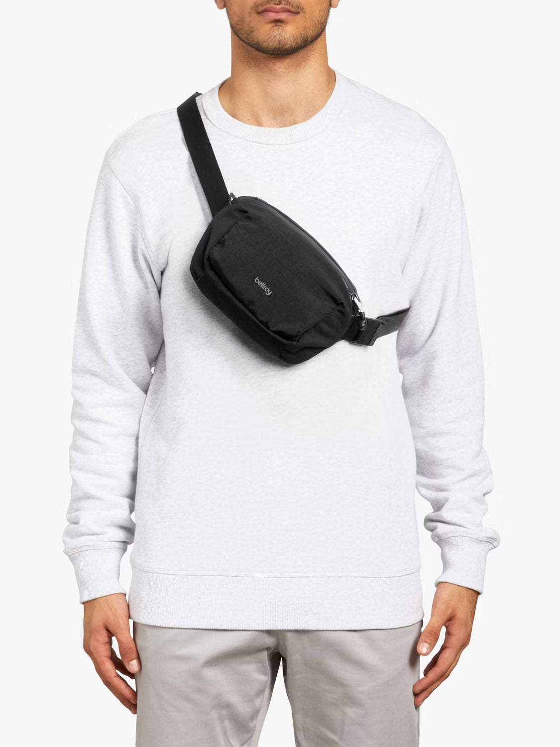 A person wears a light gray sweatshirt and beige pants, carrying the Bellroy Lite Belt Bag—a black, foldable, rectangular crossbody—over their shoulder. They stand against a plain white background.