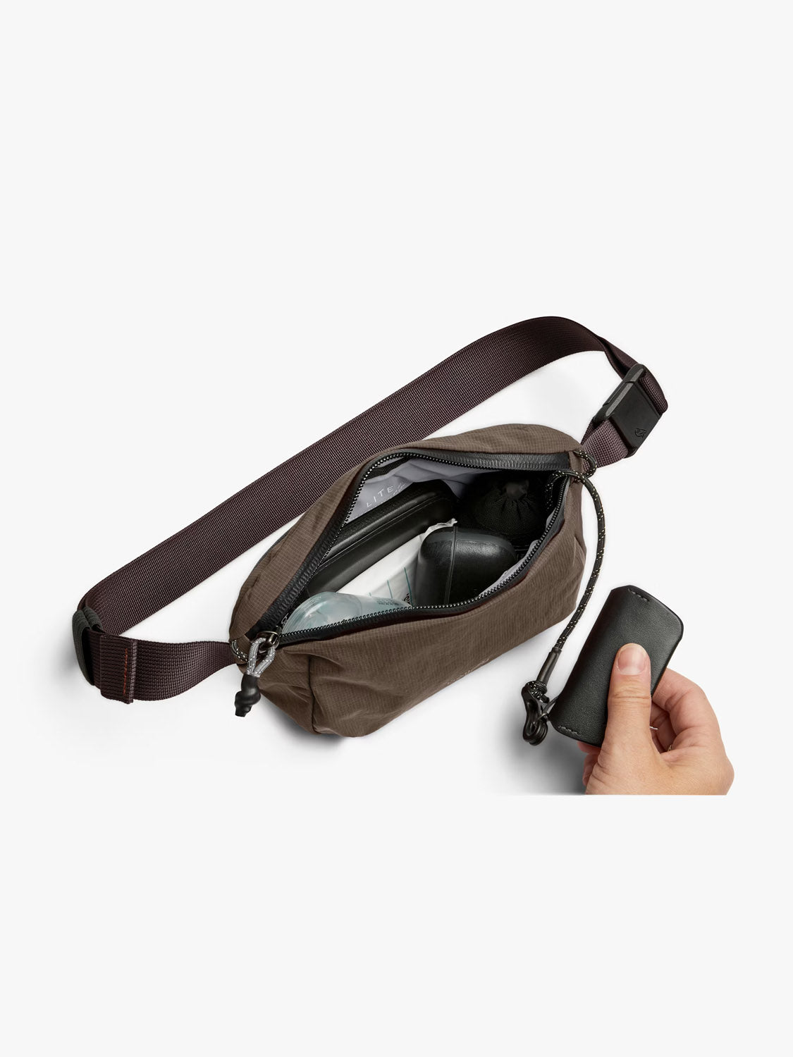 A hand holds a compact black case beside an open Bellroy Lite Belt Bag, which contains sunglasses, a pen, and other small items. The brown bag features a dark adjustable strap for easy carrying.