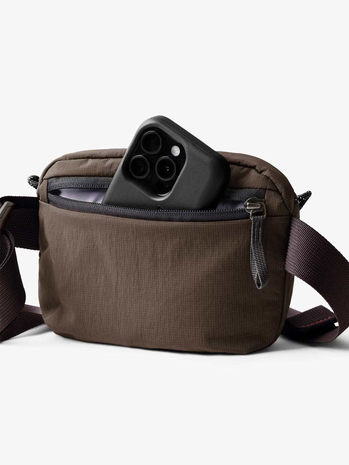 The Bellroy Lite Belt Bag is a brown, water-resistant travel bag with a dark adjustable strap and zippered front pocket. Partially open, it reveals a black smartphone with three camera lenses, all set against a white background.