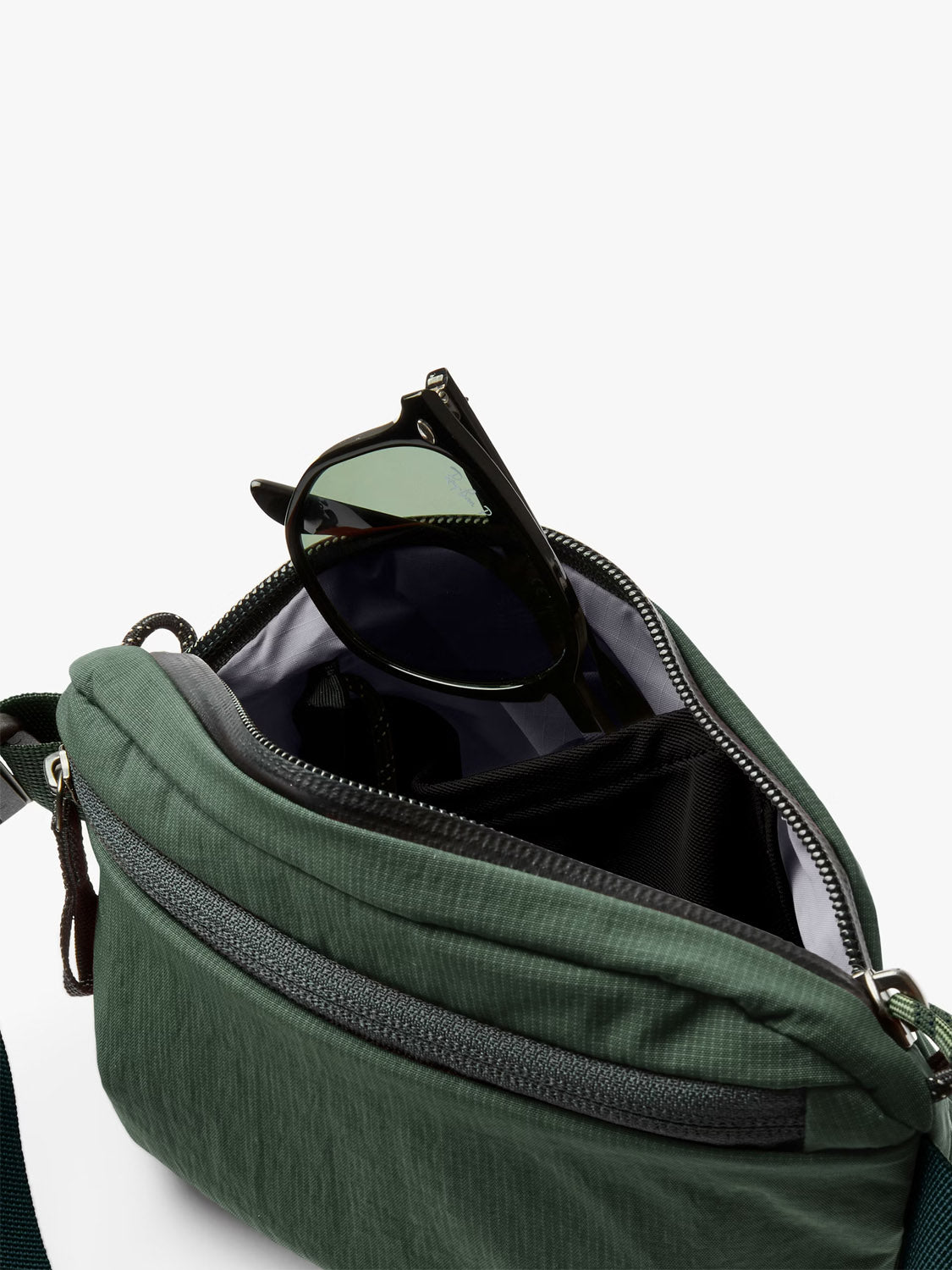 A close-up of the Bellroy Lite Belt Bag in green, featuring a black zipper and black sunglasses partially tucked inside the main compartment.