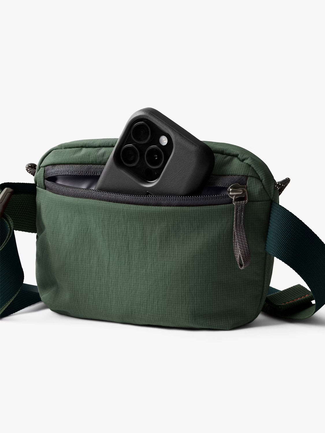 The Bellroy Lite Belt Bag is a small green, water-resistant bag with a dark zipper and adjustable strap, shown holding a black smartphone partly tucked in its front zip pocket against a plain white background.