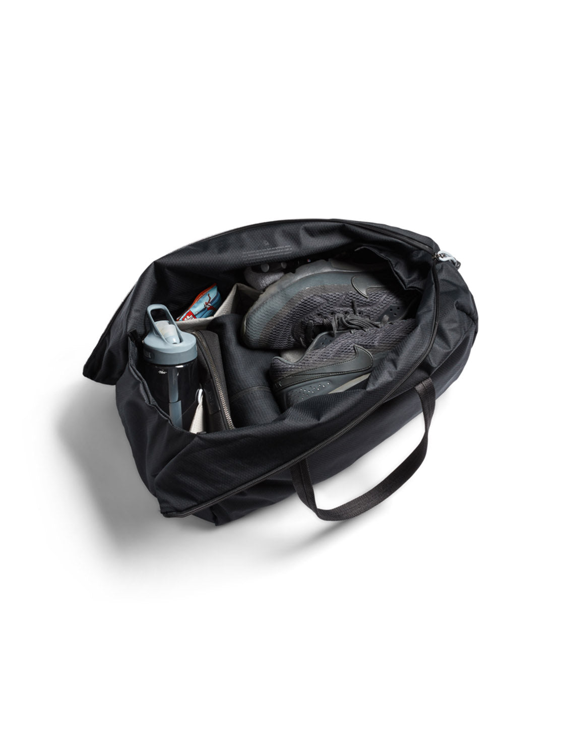 The Bellroy Lite Duffle Bag - 30L, in black and water-resistant, sits open on a white background with black athletic shoes, a rolled-up yoga mat, a water bottle, and a snack bar visible inside.