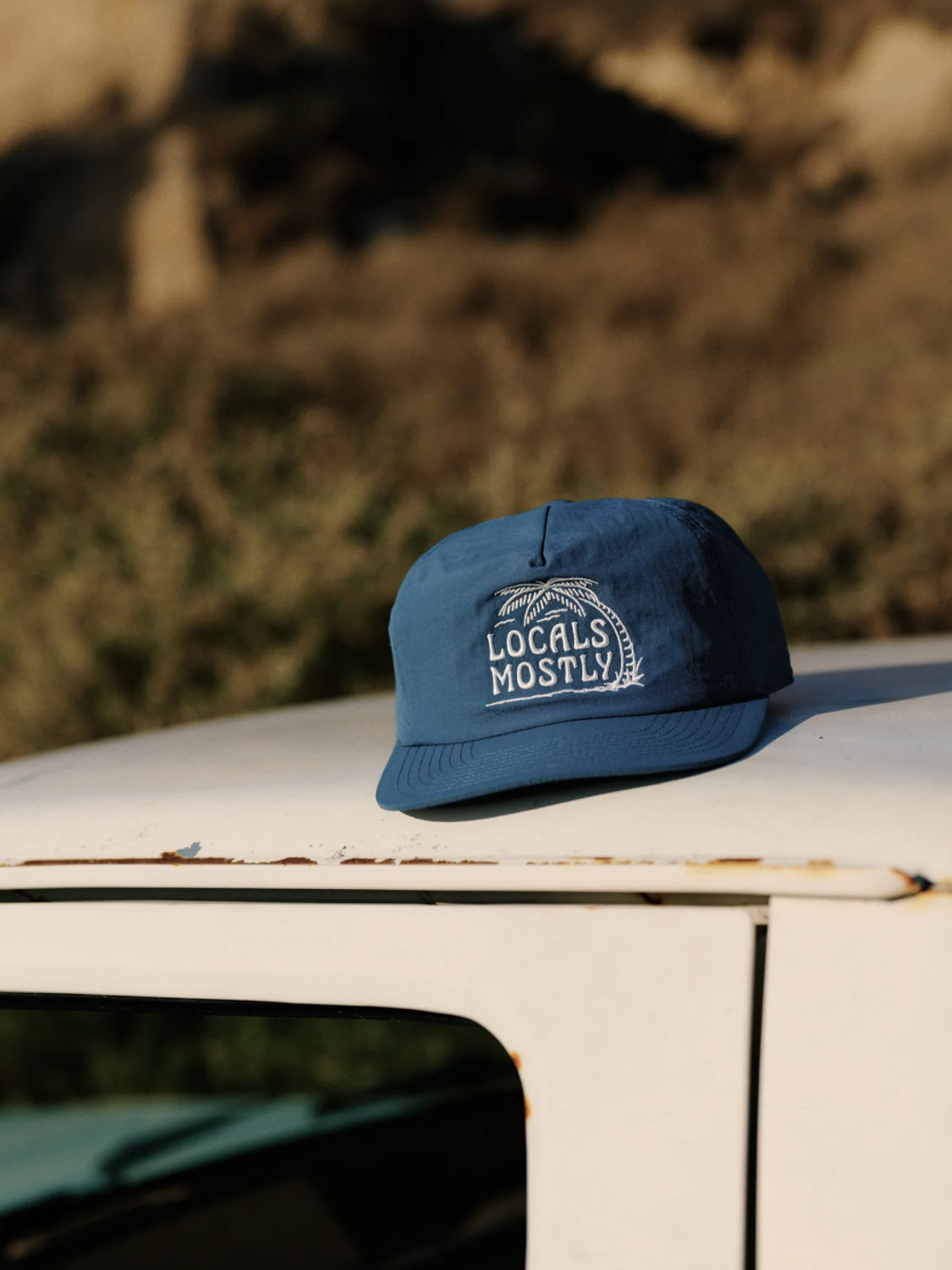 The Katin Locals Hat, a blue recycled nylon snapback with "Locals Mostly" and palm tree embroidery, sits on a white vehicle’s roof against a backdrop of blurred greenery and brown terrain.