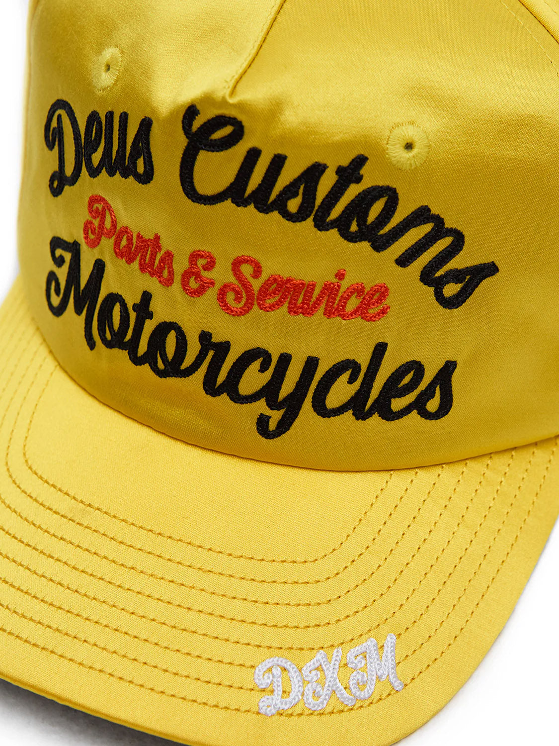 Close-up of the yellow Lota Trucker hat by Deus, featuring black and red chainstitch "Deus Customs Parts & Service Motorcycles" embroidery and white DXM initials on the bill.