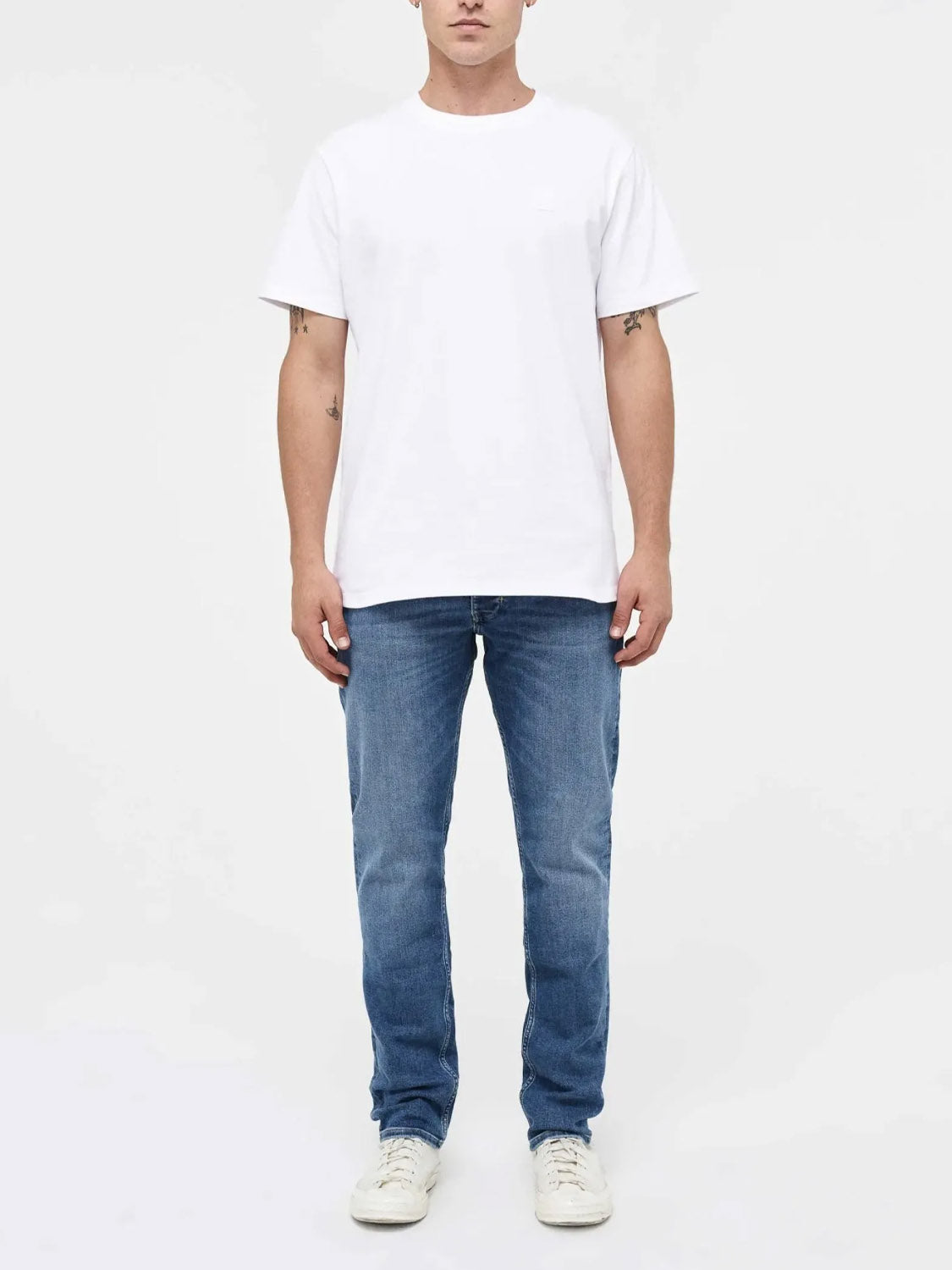 A person stands facing forward, wearing Neuw&#39;s Lou Slim Destination indigo denim jeans, a plain white organic cotton t-shirt, and white sneakers against a plain light background.
