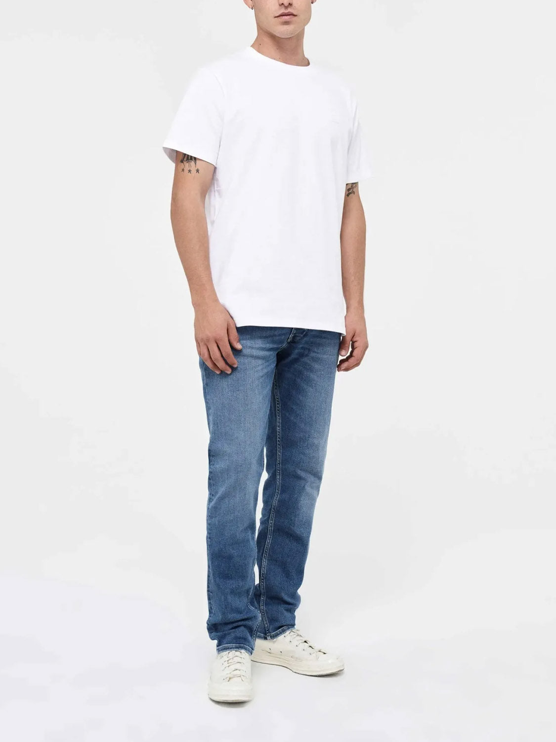 A person stands against a white background wearing Neuw's Lou Slim Destination indigo denim jeans, a white t-shirt, and white sneakers. Their face is partially out of frame.