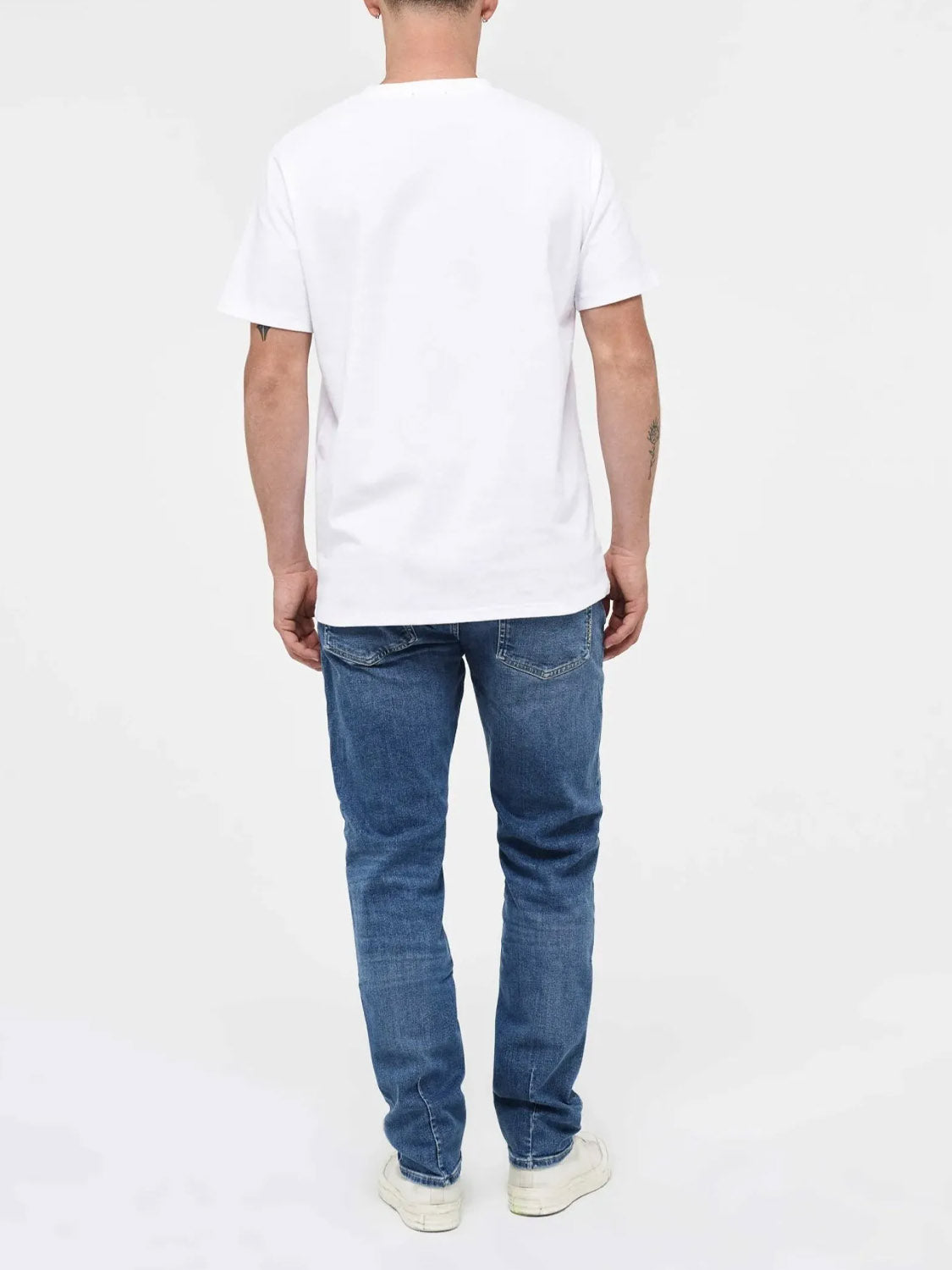 A person is facing away from the camera, wearing Neuw’s Lou Slim Destination indigo denim jeans, a plain white T-shirt, and white sneakers against a plain white background.