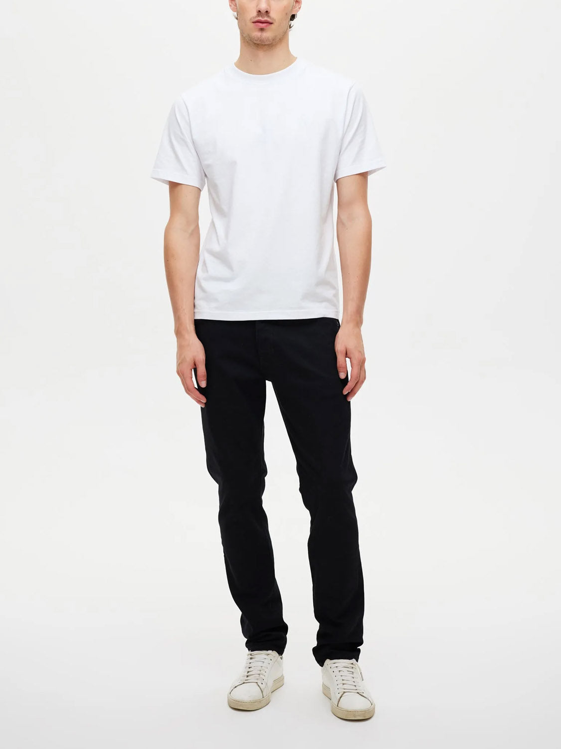 A person stands against a plain white background, wearing a white t-shirt and Neuw Lou Slim Forever Black denim slim-fit jeans with comfort stretch. Their arms are relaxed by their sides, and their face is partially visible.