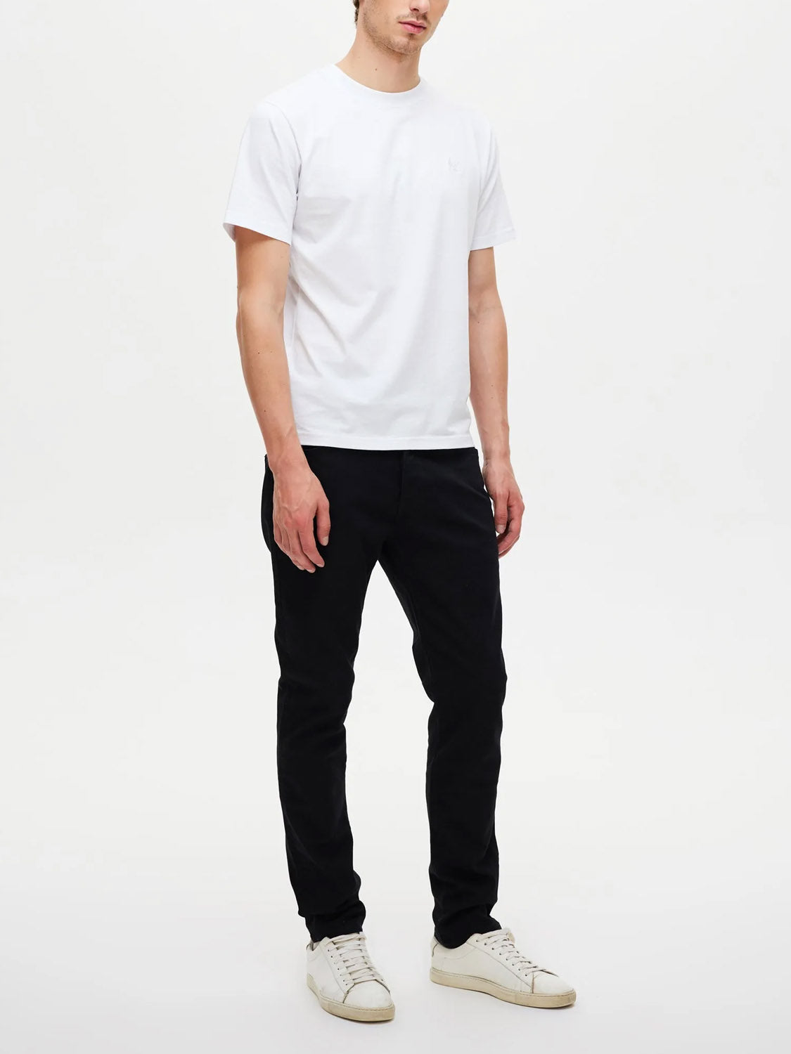 A person stands against a white background wearing a white short-sleeve t-shirt, white sneakers, and Neuw Lou Slim Forever Black comfort stretch slim-fit jeans. Their head is slightly tilted and their arms rest naturally by their sides.