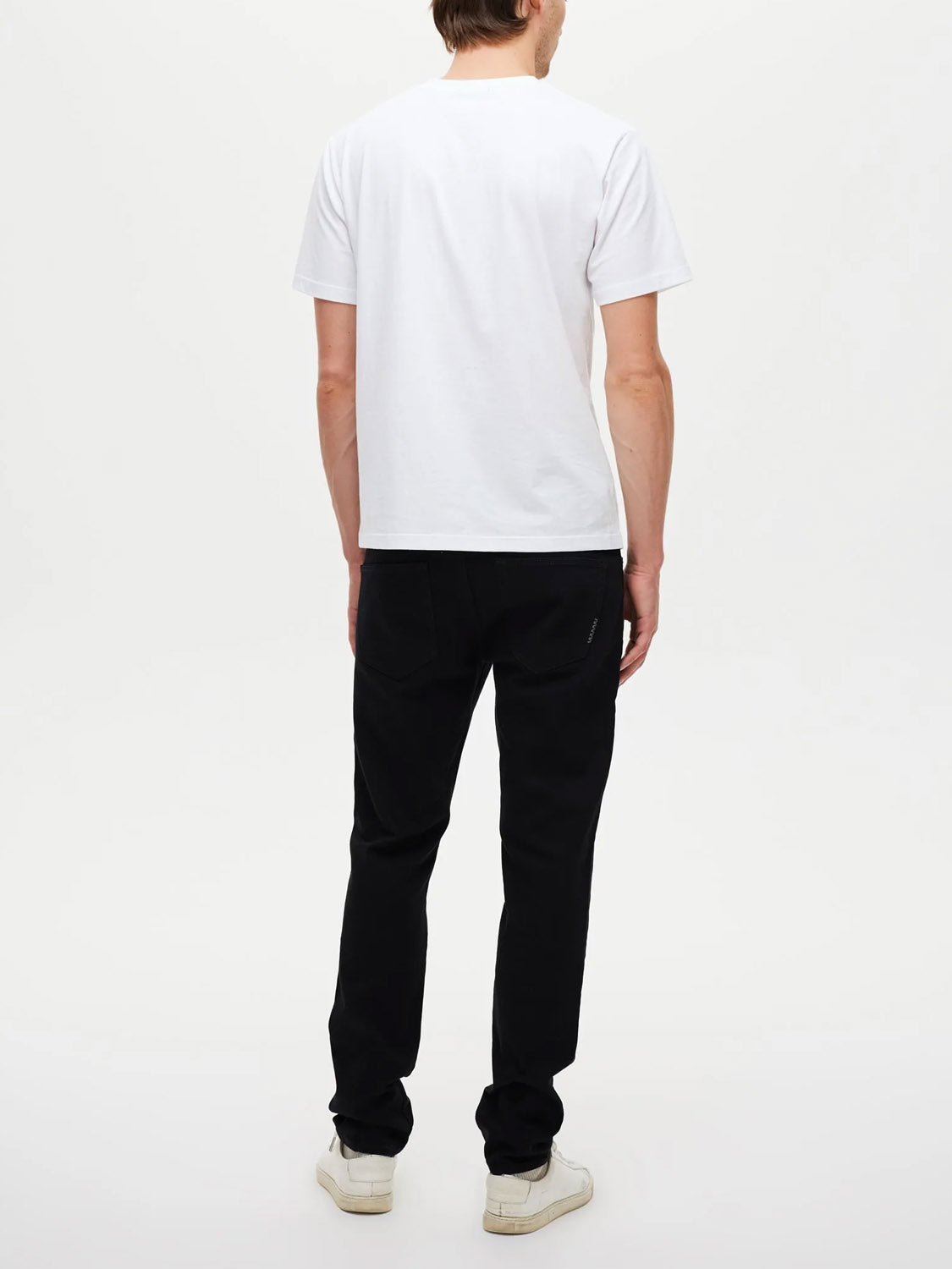 A person stands with their back to the camera, wearing Neuw's Lou Slim Forever Black jeans, a plain white short-sleeve T-shirt, and white sneakers against a plain white background.