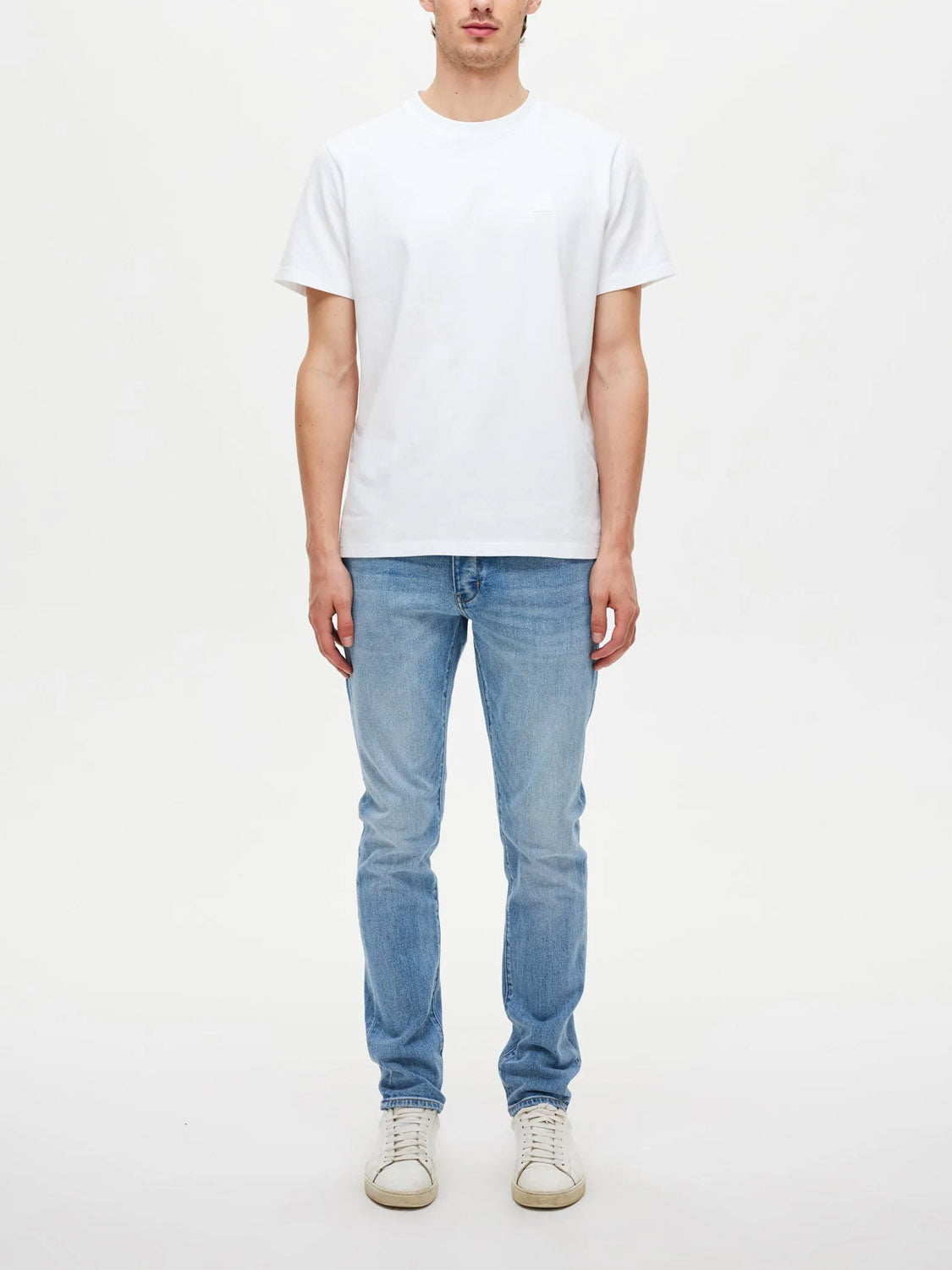 A person stands facing forward against a white background, wearing Neuw's Lou Slim Fazer slim-fit jeans in light blue with a white organic cotton t-shirt and white sneakers. Their arms are relaxed by their sides, face partially out of frame.