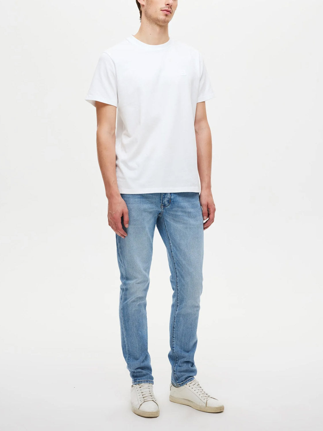 A person stands against a plain white background wearing Neuw's Lou Slim Fazer slim-fit jeans in light blue, paired with a white t-shirt and white sneakers. Their face is partially obscured, and their arms hang relaxed by their sides.