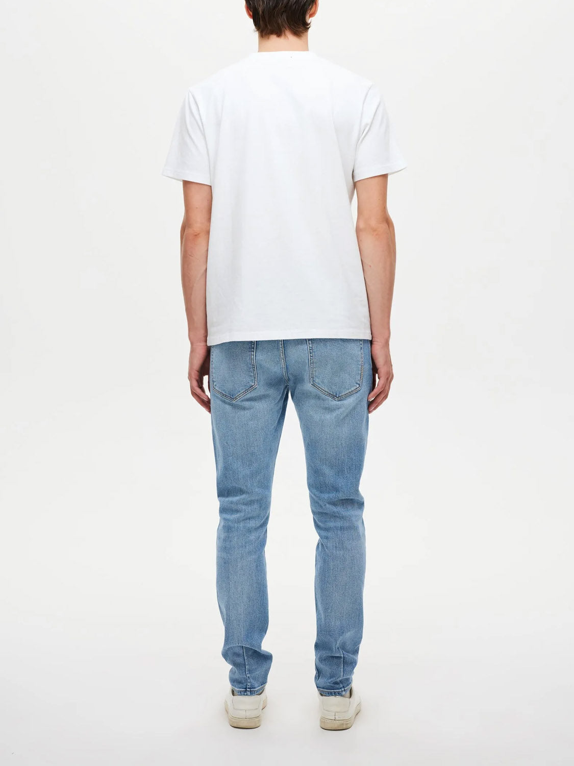 A person stands facing away from the camera, wearing Neuw's Lou Slim Fazer jeans in light blue indigo denim, a plain white t-shirt, and white sneakers against a plain white background.