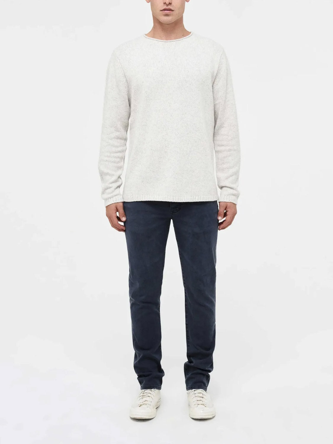 A person stands facing forward against a plain white background, wearing Neuw’s Lou Slim Liberte dark blue slim-fit jeans with a light beige sweater and white sneakers. The top of their head is partially cropped out of the image.