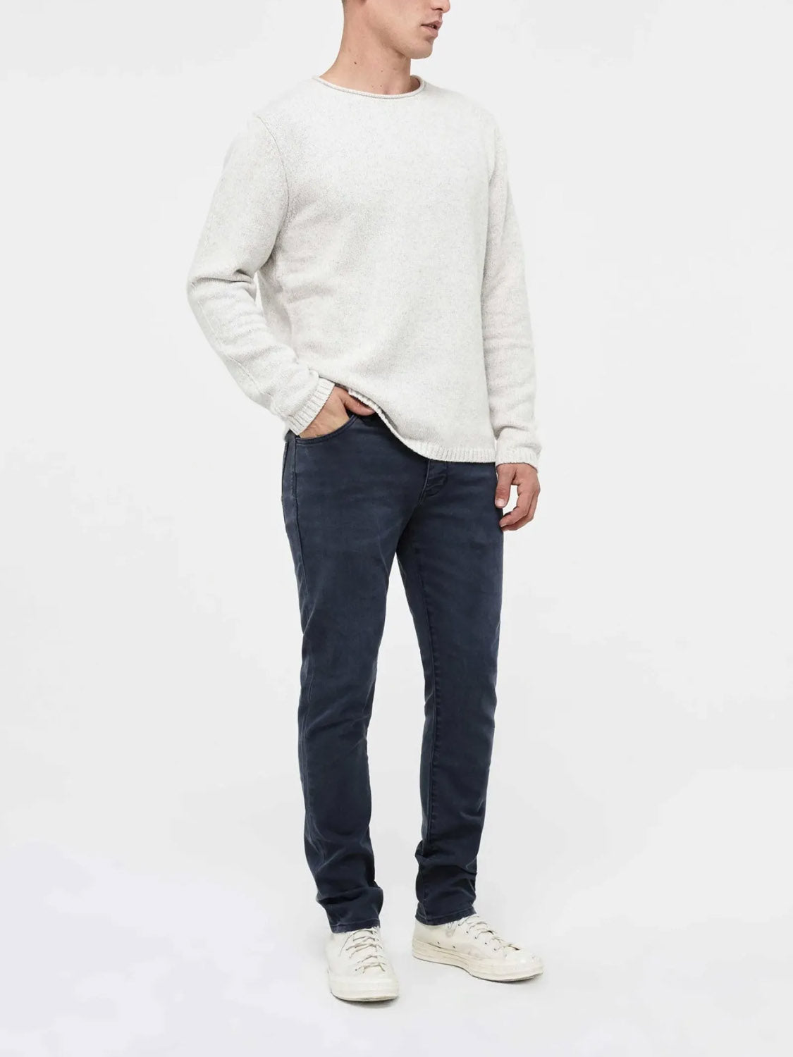 A person stands against a plain light background, wearing the Neuw Lou Slim Liberte dark blue straight-leg jeans with premium stretch denim, paired with a light gray sweater and white sneakers. Their head is turned slightly and their right hand rests in their pocket.