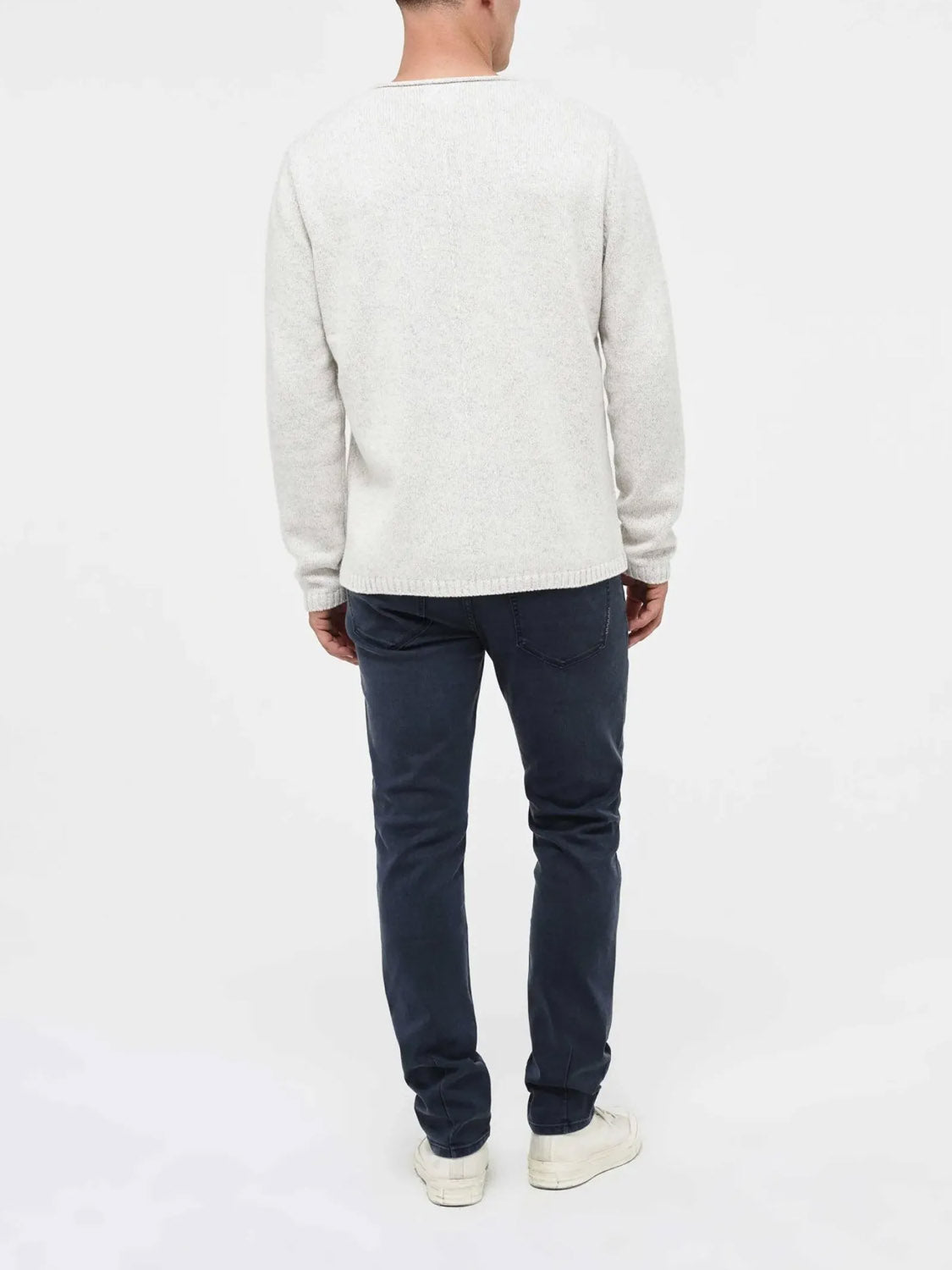 A person stands with their back to the camera, wearing a light gray sweater, white sneakers, and Neuw’s Lou Slim Liberte premium stretch denim jeans in dark blue against a plain white background.
