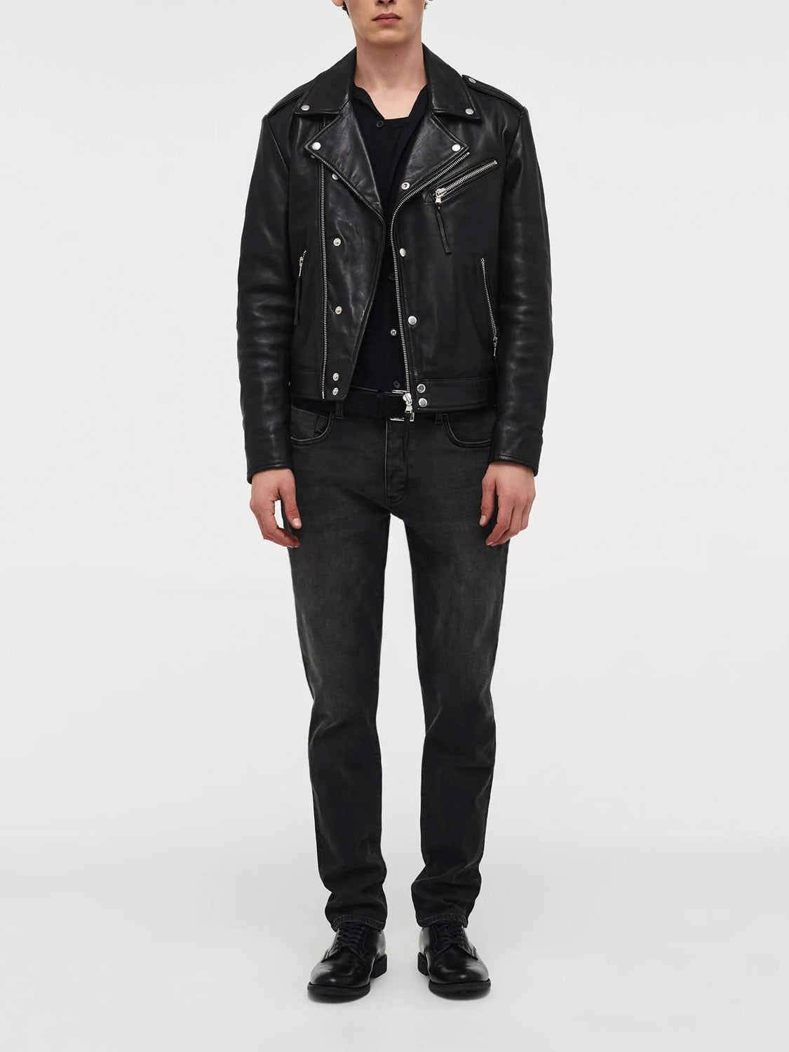 A person stands against a plain background wearing a black leather jacket, black shirt, Neuw&#39;s Lou Slim Moonshake premium denim jeans, and black shoes, with their arms relaxed at their sides.