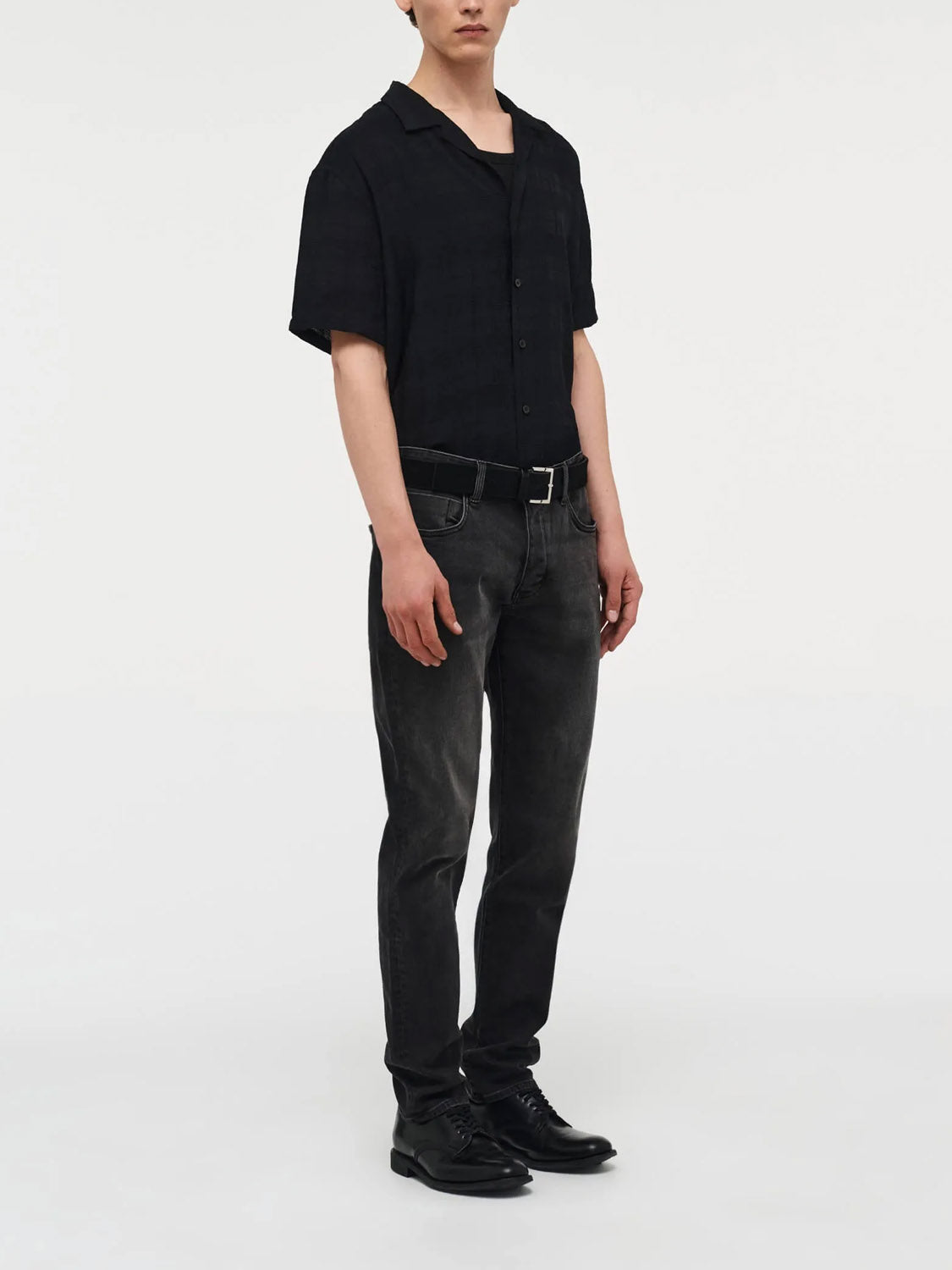 A person stands against a white background wearing Neuw's Lou Slim Moonshake jeans with a black short-sleeved button-up, a belt, and black shoes. Their pose is relaxed with hands by their sides.