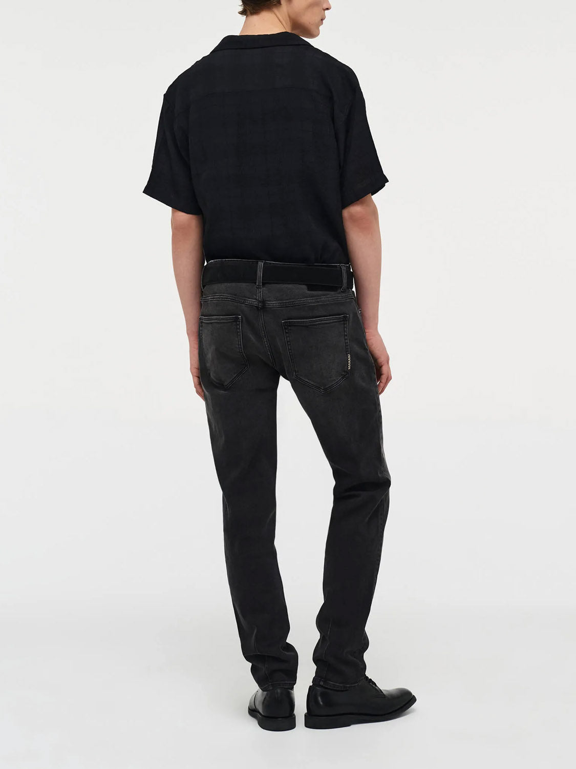 A person faces away from the camera, wearing Neuw's Lou Slim Moonshake jeans in faded black with a short-sleeve black shirt and black shoes, set against a plain white background.