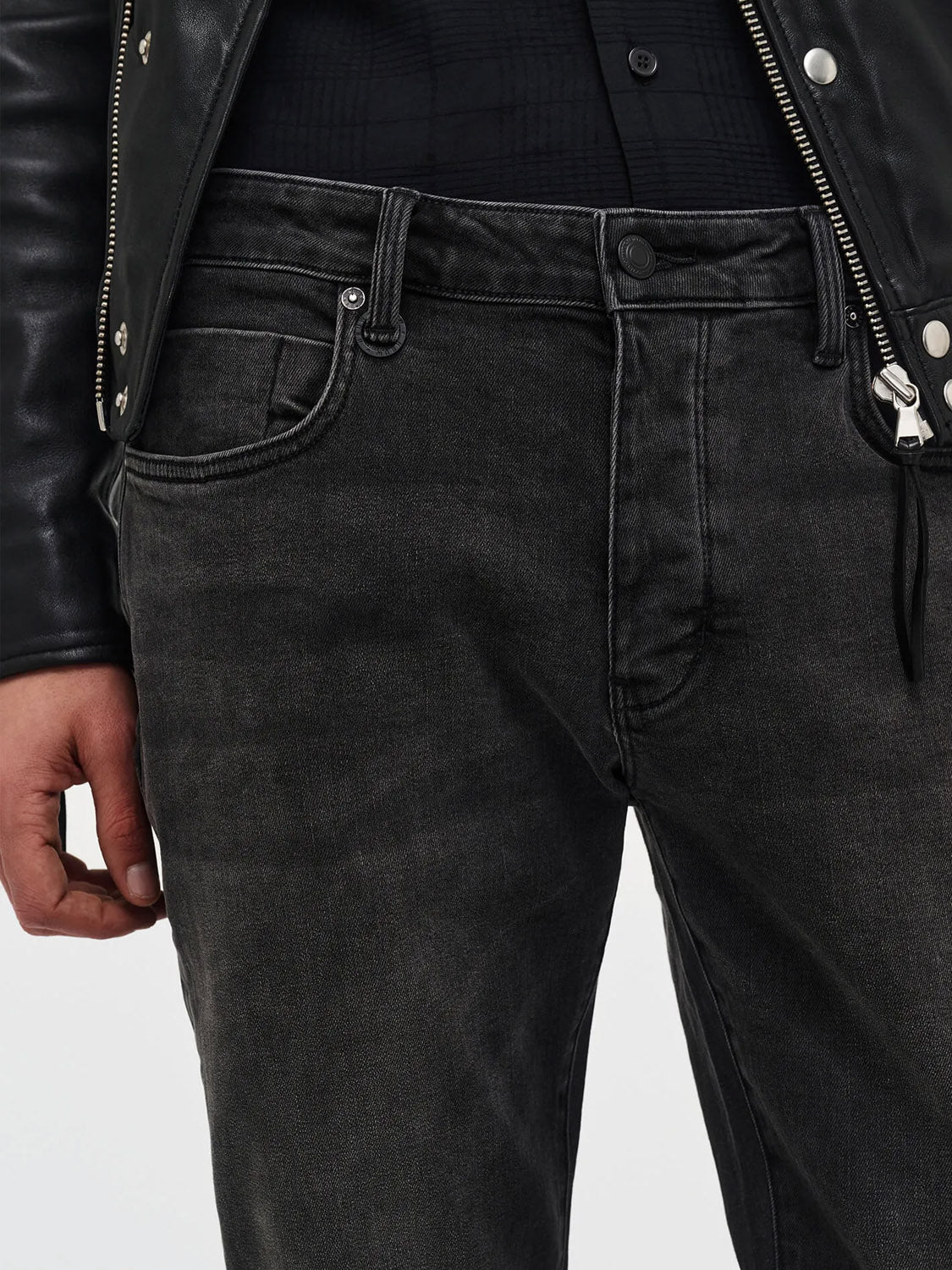 Close-up of a person wearing Neuw's Lou Slim Moonshake jeans—faded black, slim fit—paired with a black leather jacket. The shot shows the torso and upper legs, with the left hand relaxed at the side.