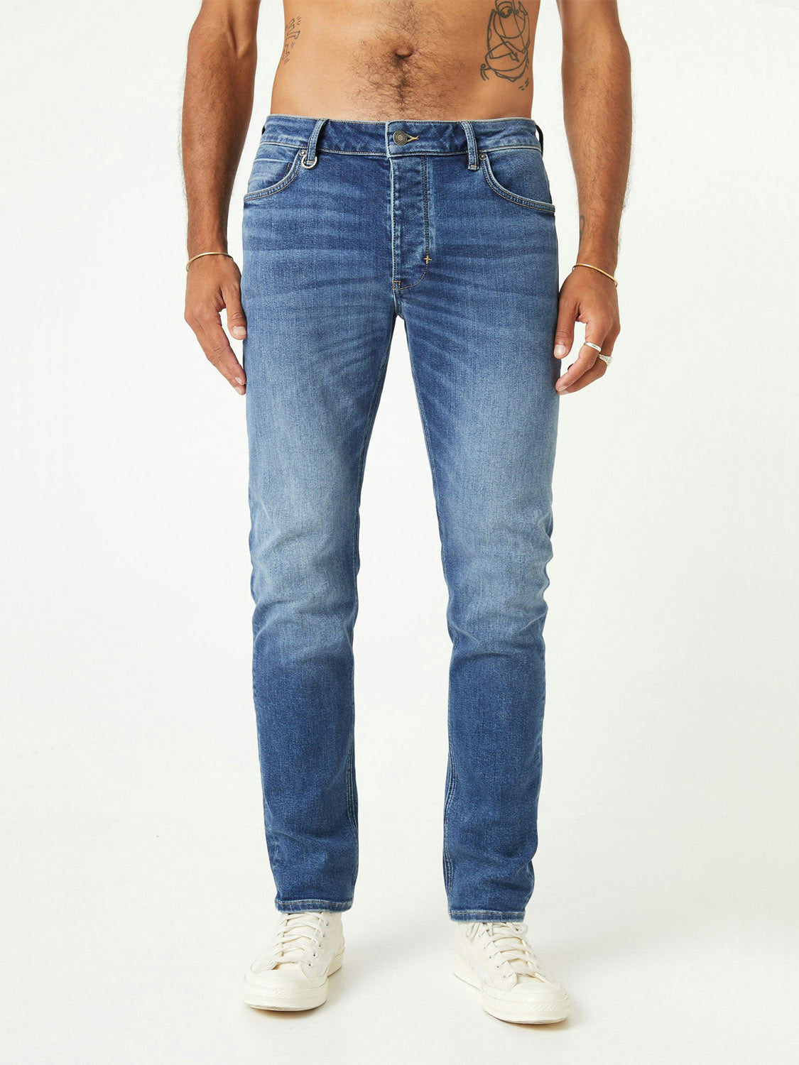 A man wearing Neuw Lou Slim Destination jeans and white sneakers stands against a white background, shown from torso to feet, with his hands by his sides.