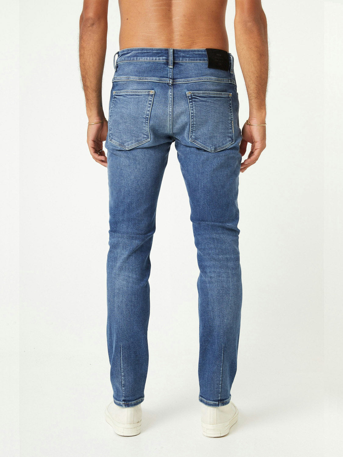 A shirtless person, seen from the back, wears Neuw Lou Slim Destination light blue jeans made from organic denim and white sneakers, standing against a plain white background.