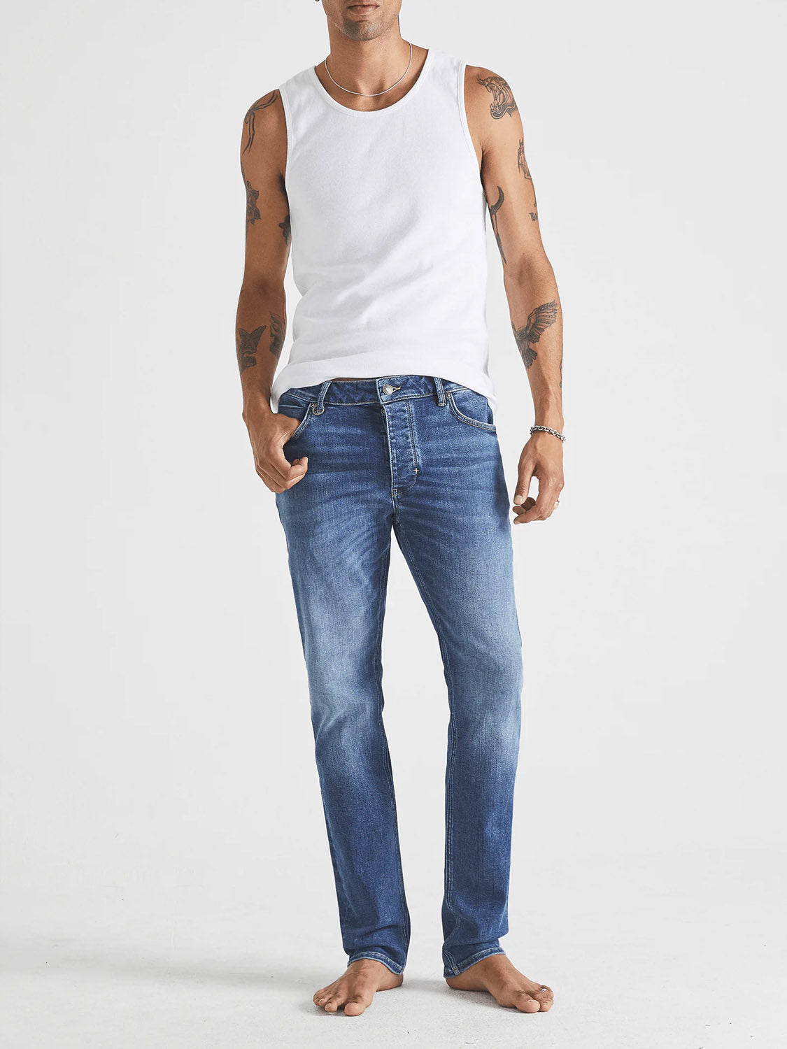 A tattooed man wears a white tank top and Neuw Lou Slim Destination jeans, standing barefoot against a plain white background.