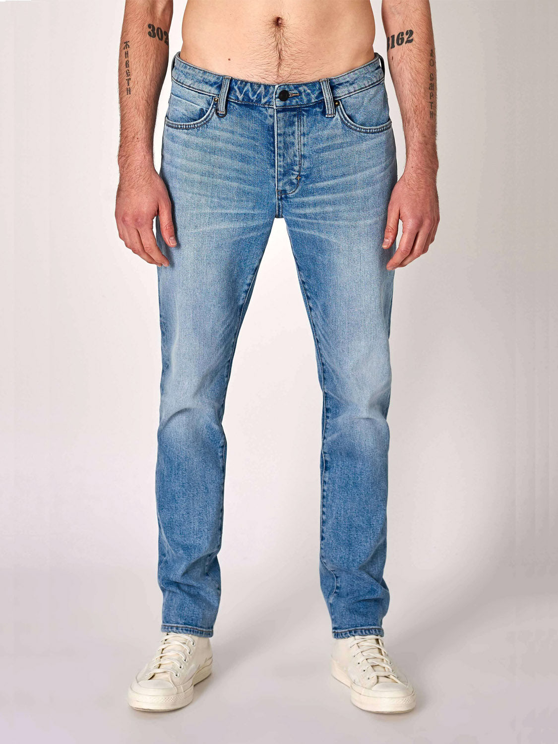 A person wearing Neuw Lou Slim Fazer jeans stands against a plain light background. The photo, cropped from shoulders down, shows only the torso, legs, and feet, with the jeans paired with white sneakers.