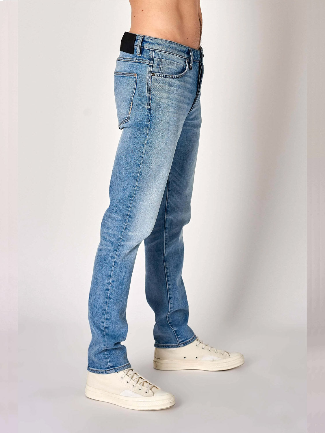 A person is shown from the waist down wearing Neuw Lou Slim Fazer light blue organic cotton denim jeans with white sneakers, standing sideways against a plain background.
