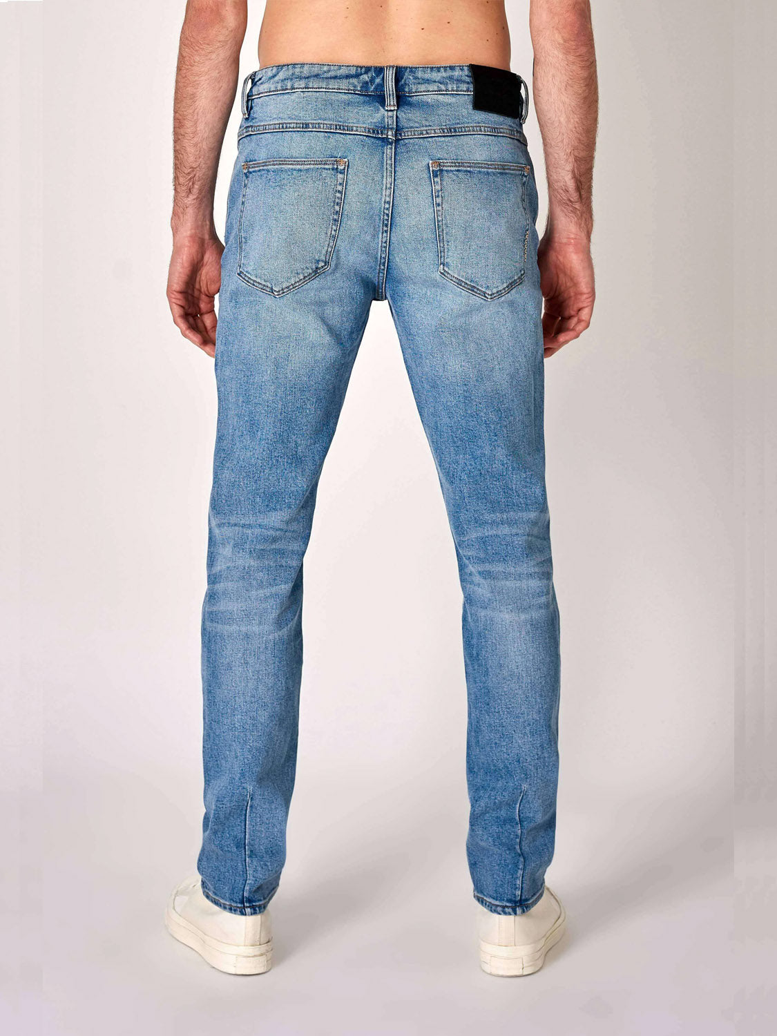 A person is seen from behind wearing Neuw's Lou Slim Fazer jeans, made from organic cotton denim, paired with white sneakers, showing the slim fit's back view against a plain backdrop.