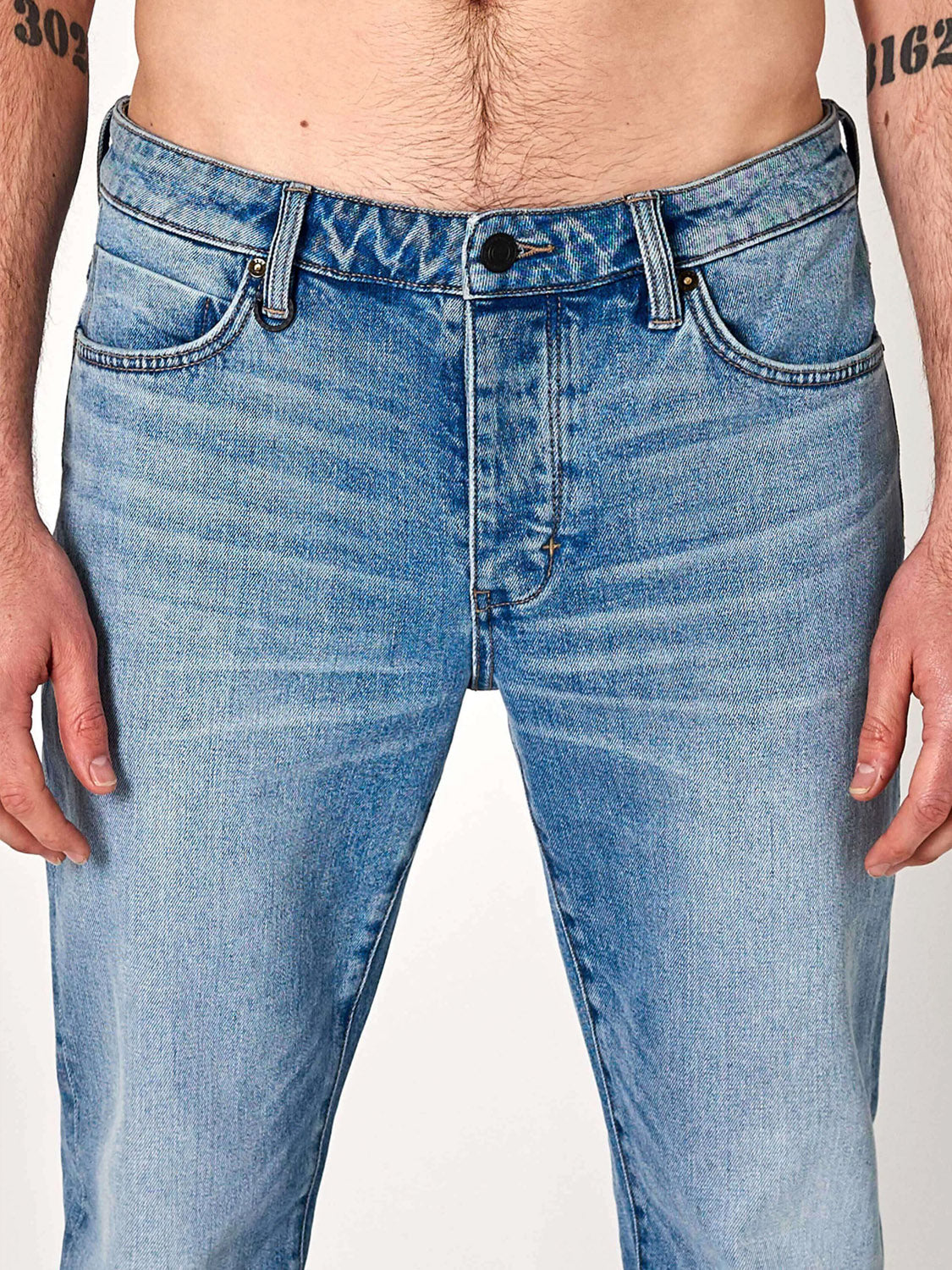 A model wears Neuw's Lou Slim Fazer light blue jeans, crafted from organic cotton denim, shown from waist to mid-thigh with a bare torso and forearm tattoos against a plain white background.