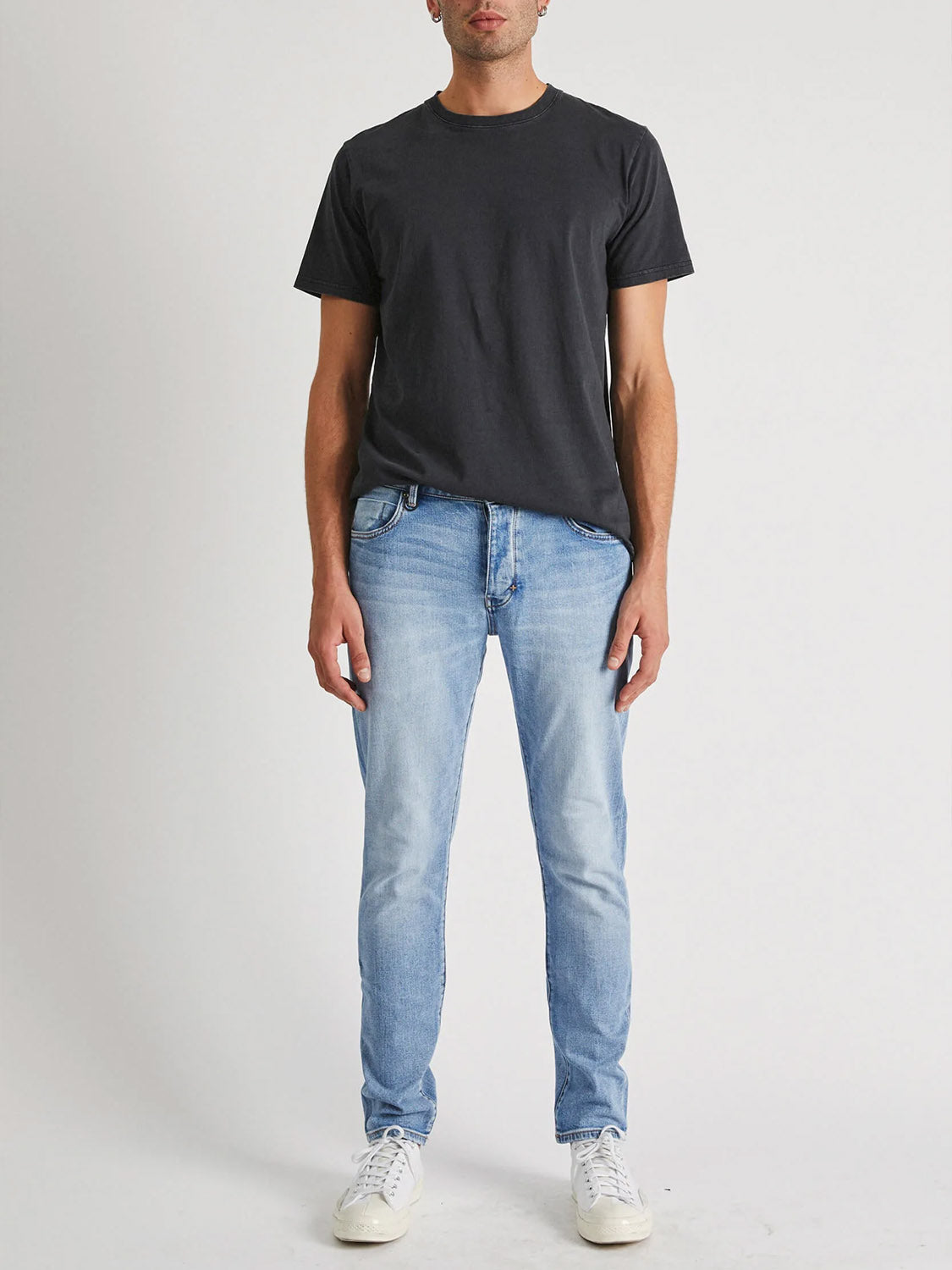 A man stands against a white background wearing a black t-shirt, Neuw Lou Slim Fazer jeans in organic cotton denim, and white sneakers. His arms are relaxed at his sides and his face is partly out of frame.