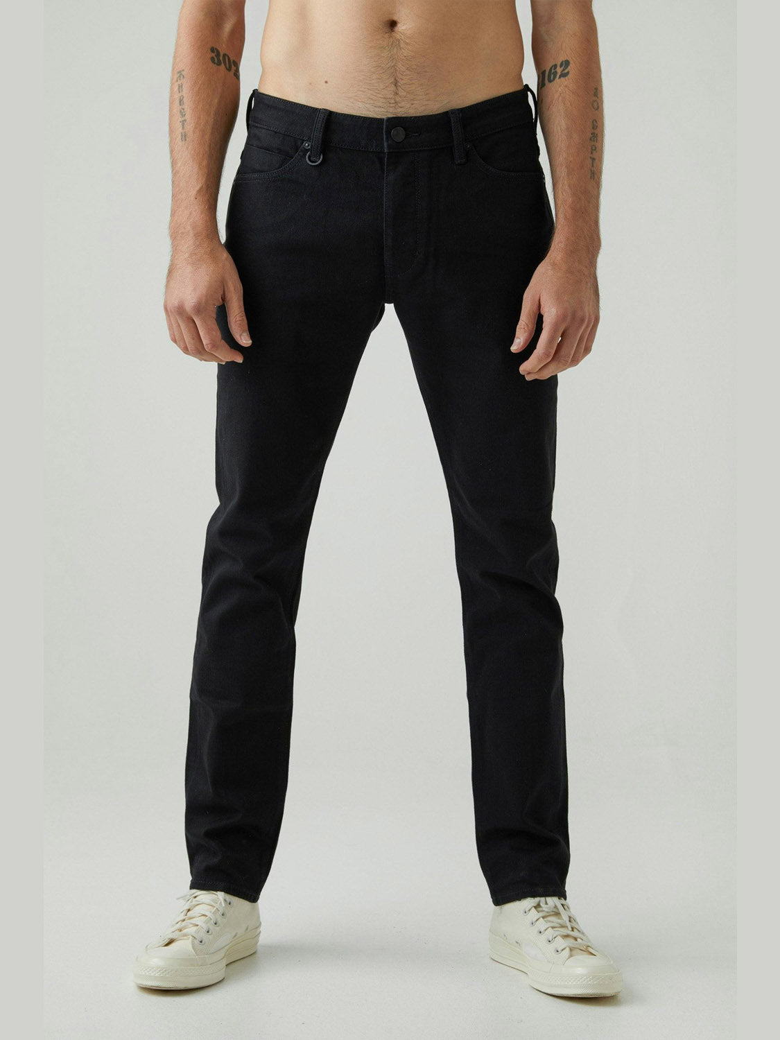 A man faces forward wearing Neuw's Lou Slim Forever Black jeans with white sneakers. His torso is bare, displaying forearm tattoos. The backdrop is plain and light gray.